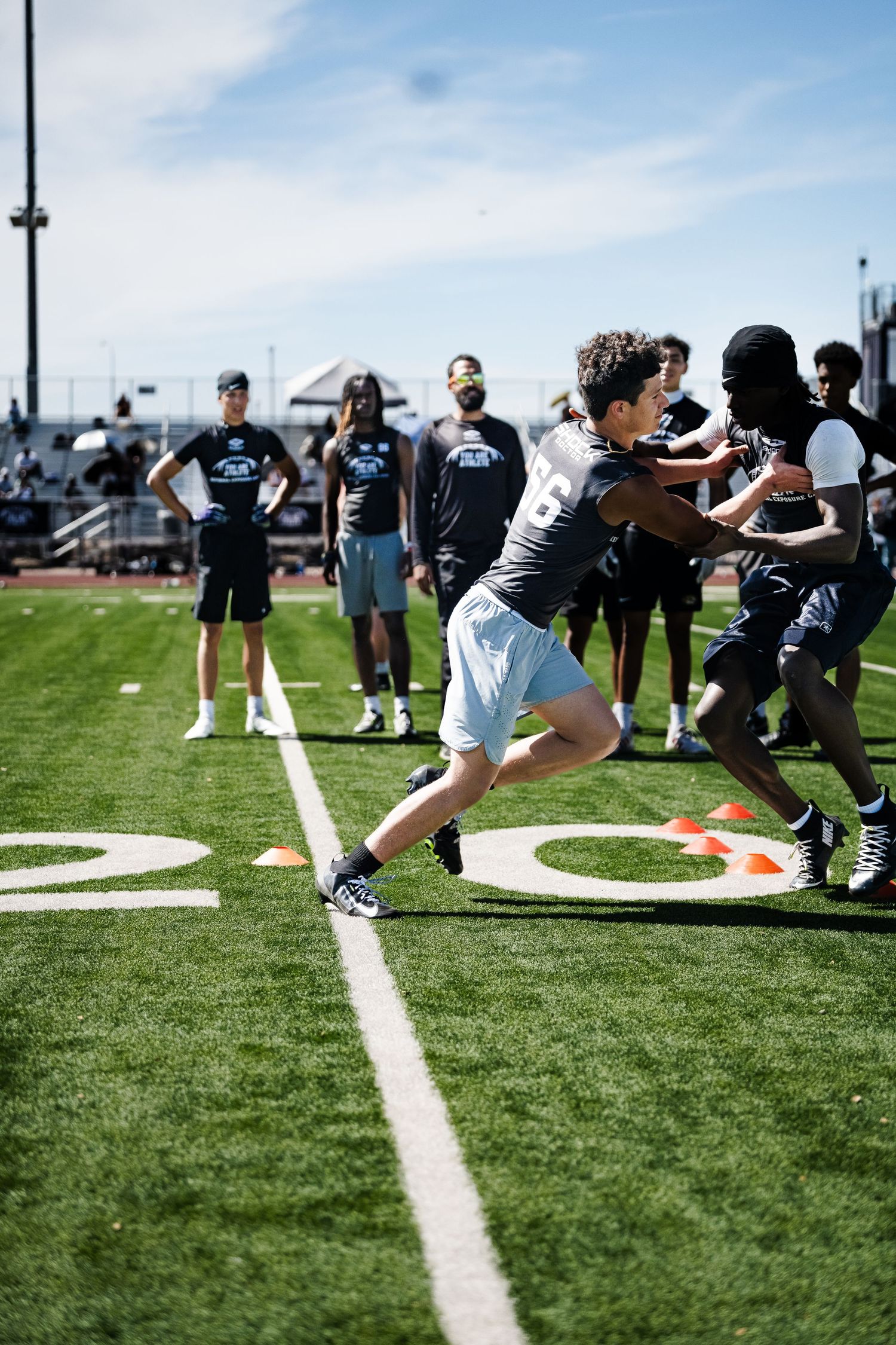 Young football players practice one-on-one blocking drills while coaches observe at an outdoor camp.