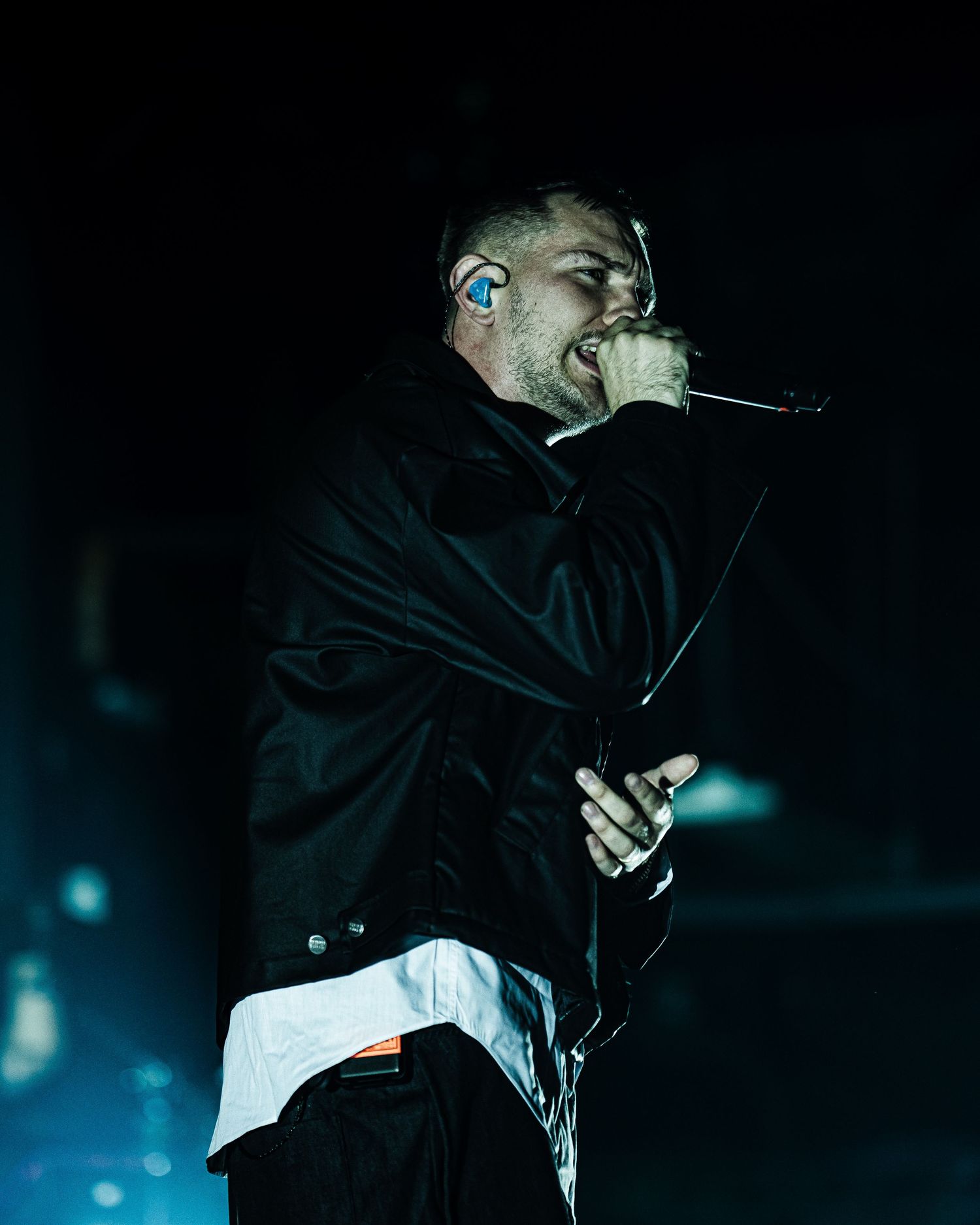 Male singer performing on dark stage, wearing black jacket, holding microphone, blue in-ear monitor visible.