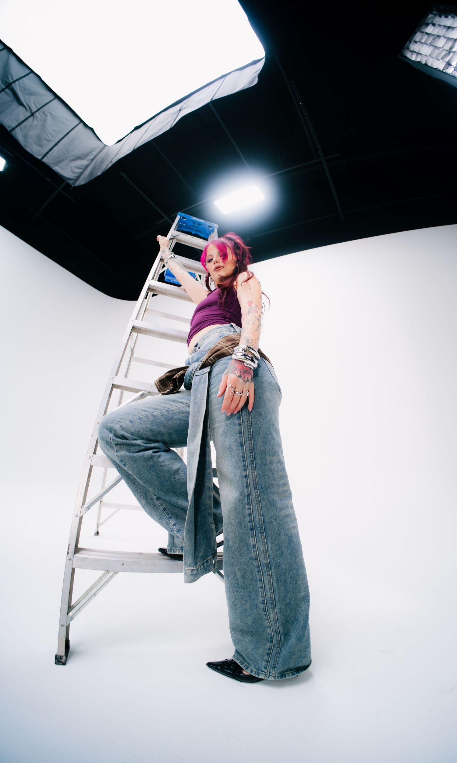 Woman with pink hair poses on a white aluminum ladder in a photography studio wearing wide-leg jeans and purple top.