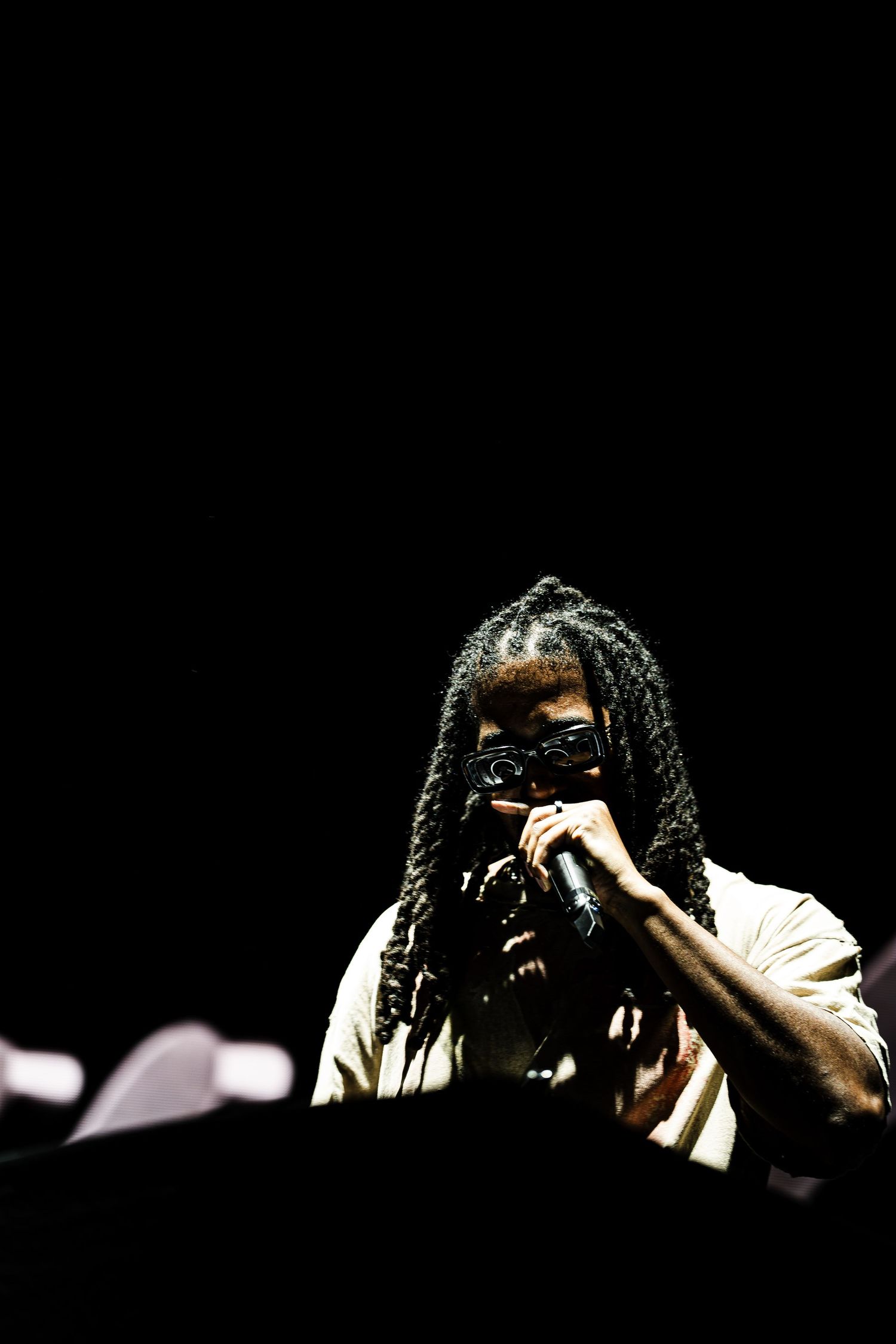 Performer with long braids and dark sunglasses holds a microphone to their mouth on a dark stage, wearing a light shirt behind equipment.