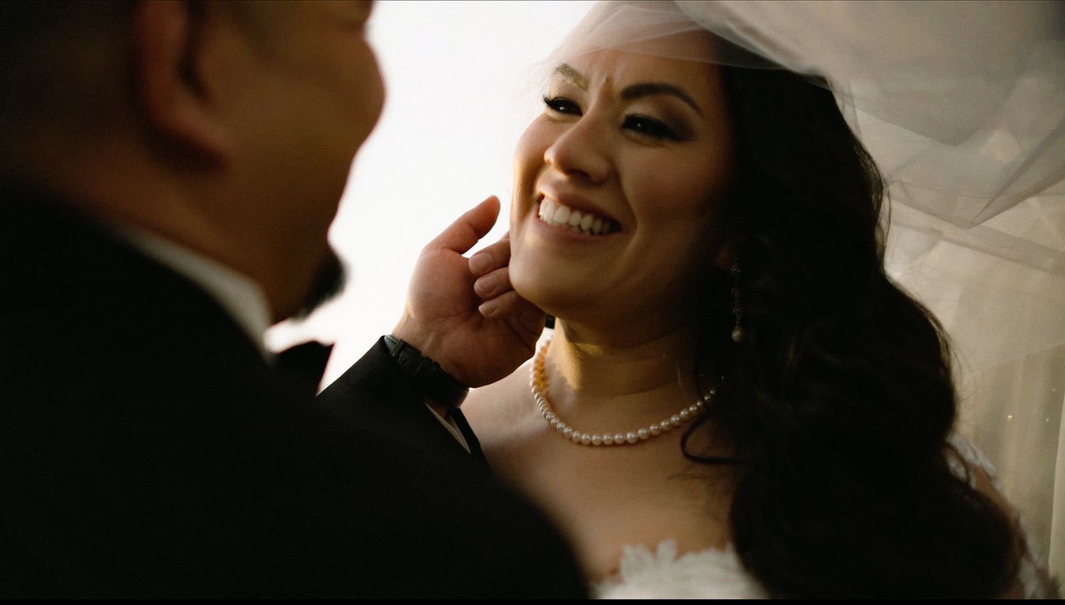A bride in a white gown and veil smiles as the groom gently touches her face during their wedding ceremony.