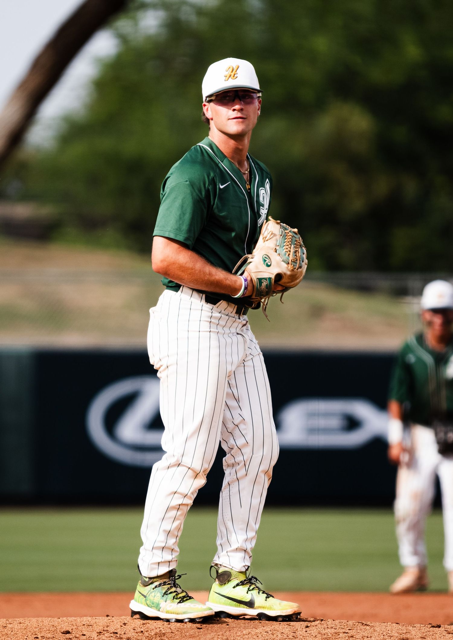 Baseball player in green and white pinstripe uniform stands on the mound holding glove during a college game.