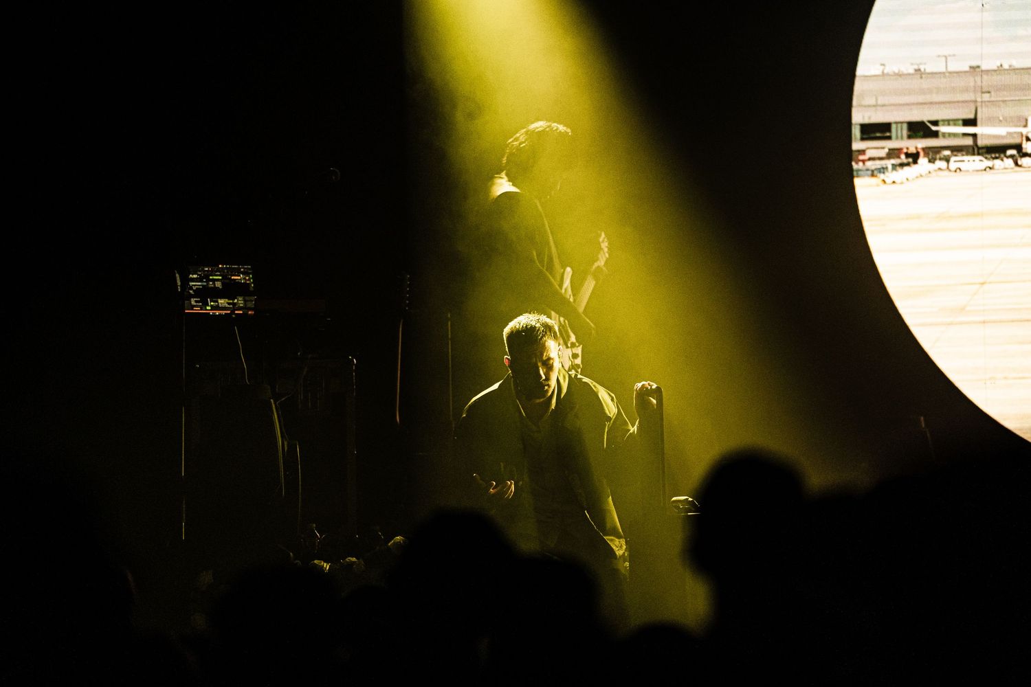 Band performing on stage under dramatic yellow spotlights with silhouetted crowd in foreground at live concert.