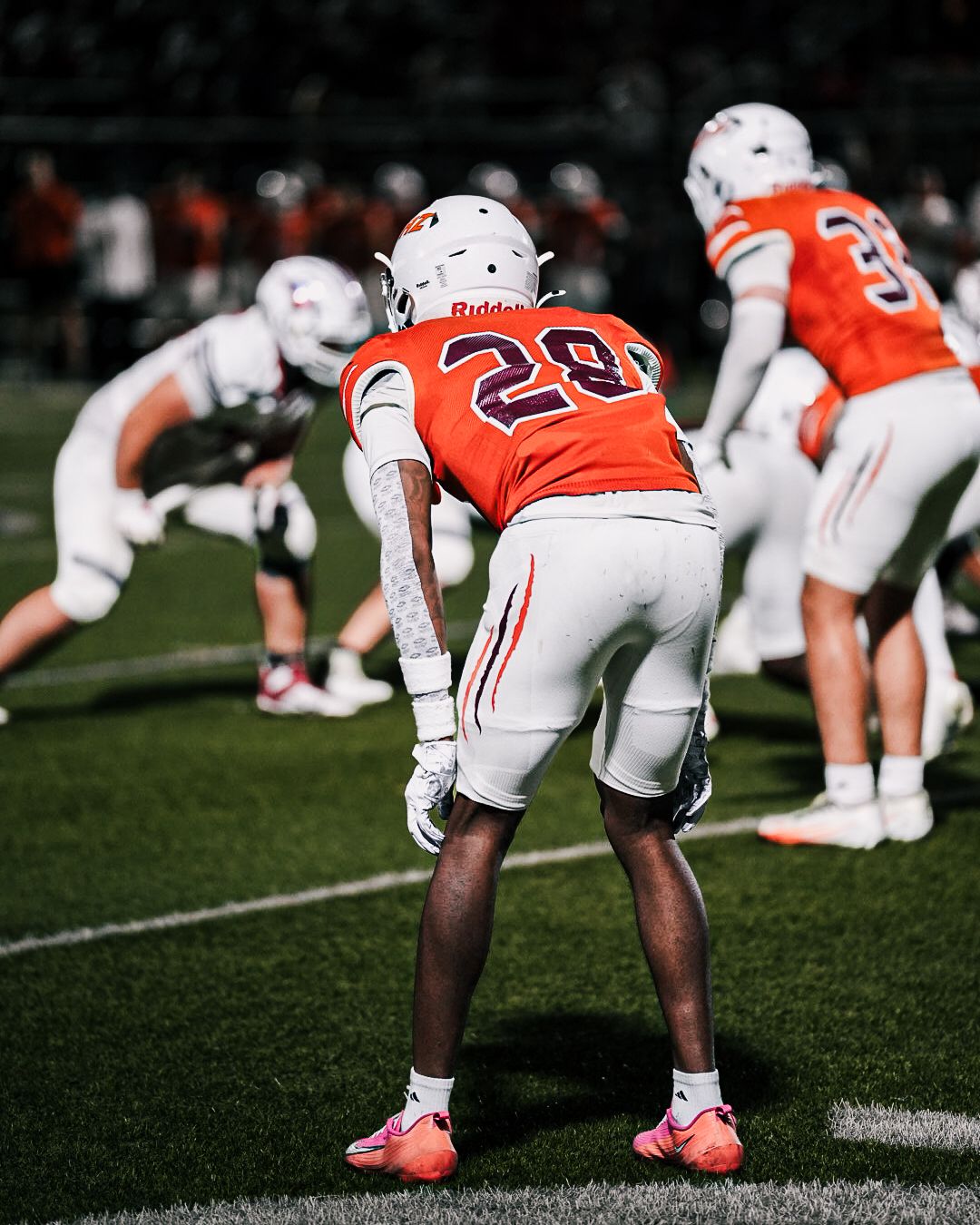 Football player wearing orange jersey number 28 stands on field during a night game, facing the line of scrimmage.
