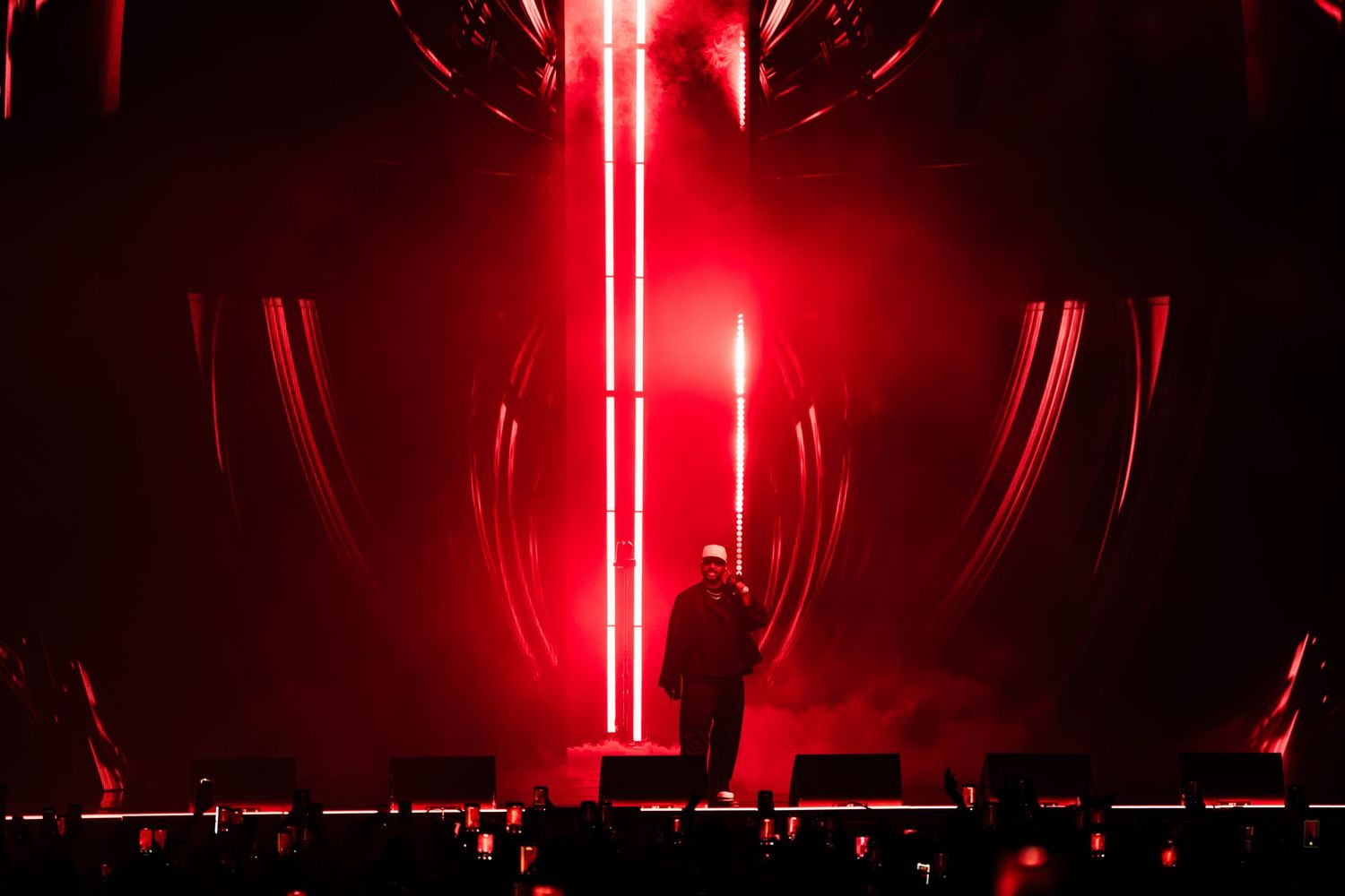 Lone performer stands on a dark concert stage bathed in intense red light, with glowing vertical beams and curved light arcs behind.