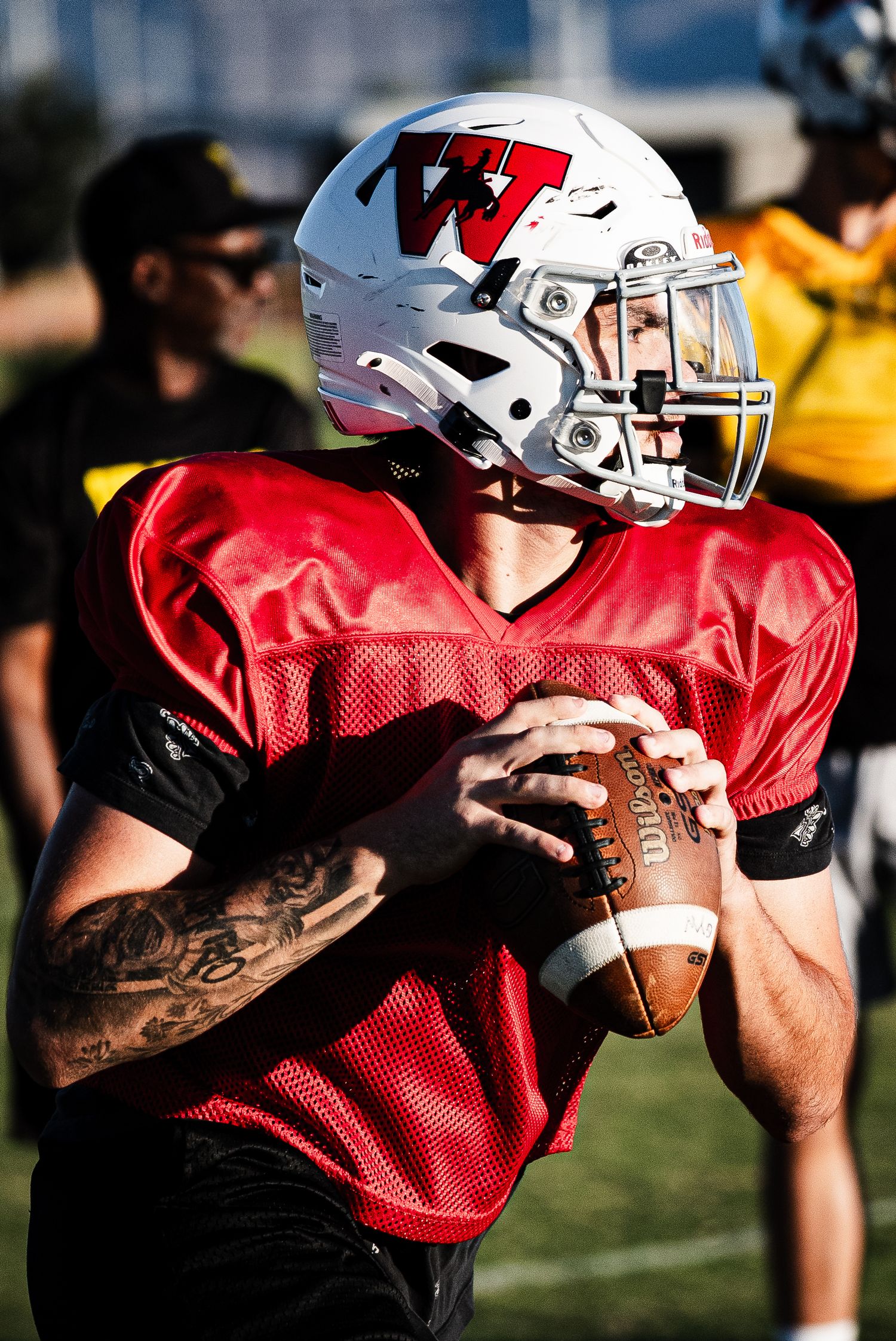A tattooed quarterback in a red jersey and white helmet holds a football in throwing position during practice.