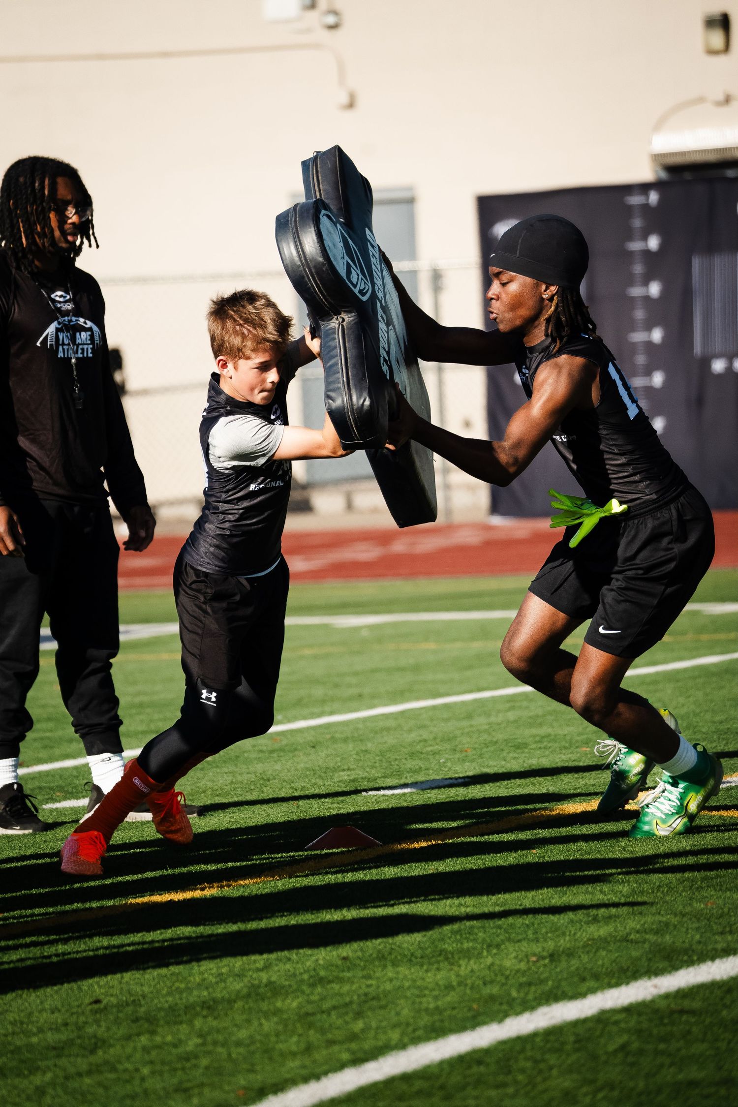 Two young football players competing in blocking drill with pad on sunny turf field.