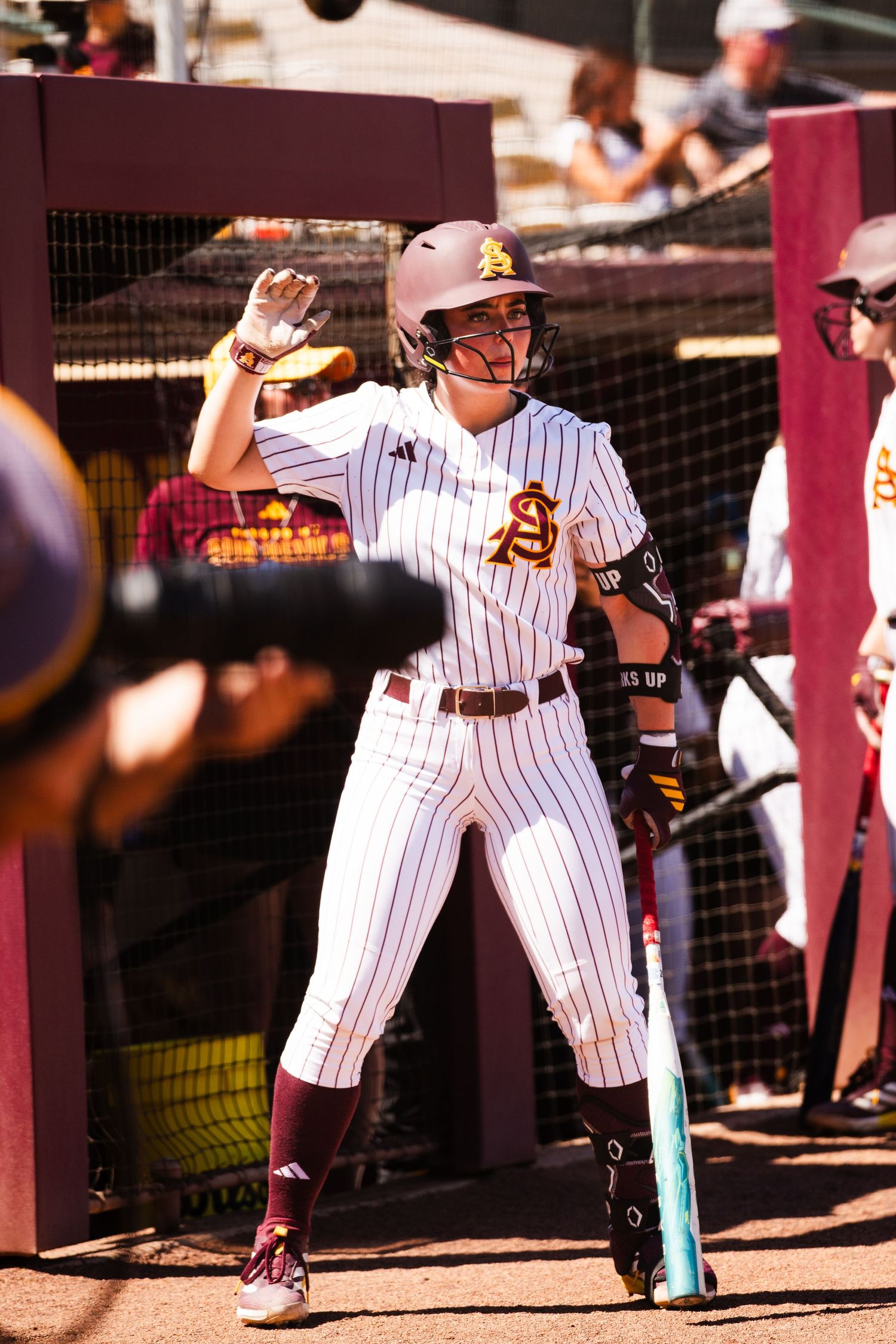 Arizona State Sun Devils softball player in white pinstripe uniform stands ready with bat at home plate during a sunny game.