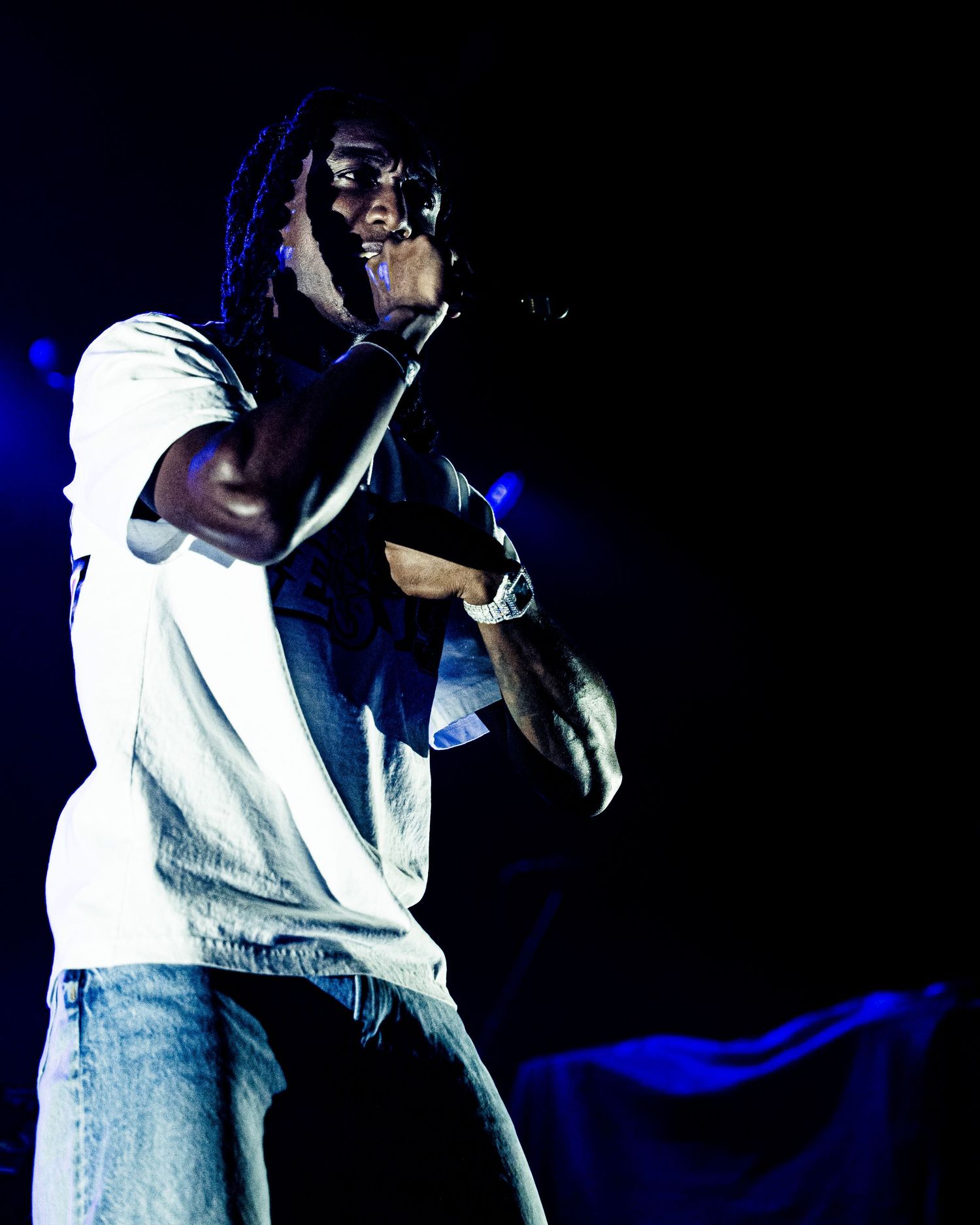 Performer with dreadlocks singing into a microphone on a dark stage with blue lighting.