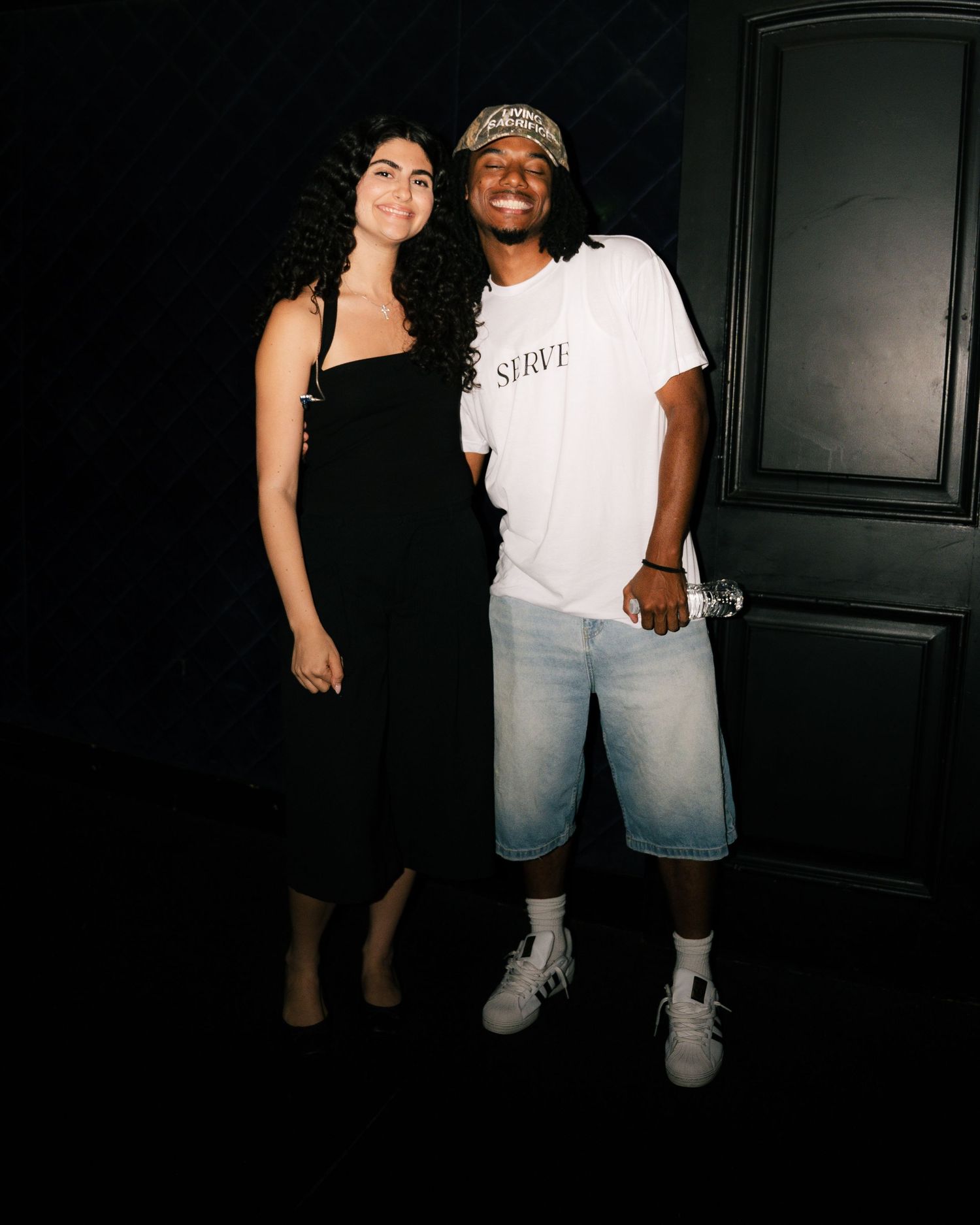 Two friends posing together, woman in black dress and man in white tee and denim shorts, against dark background.