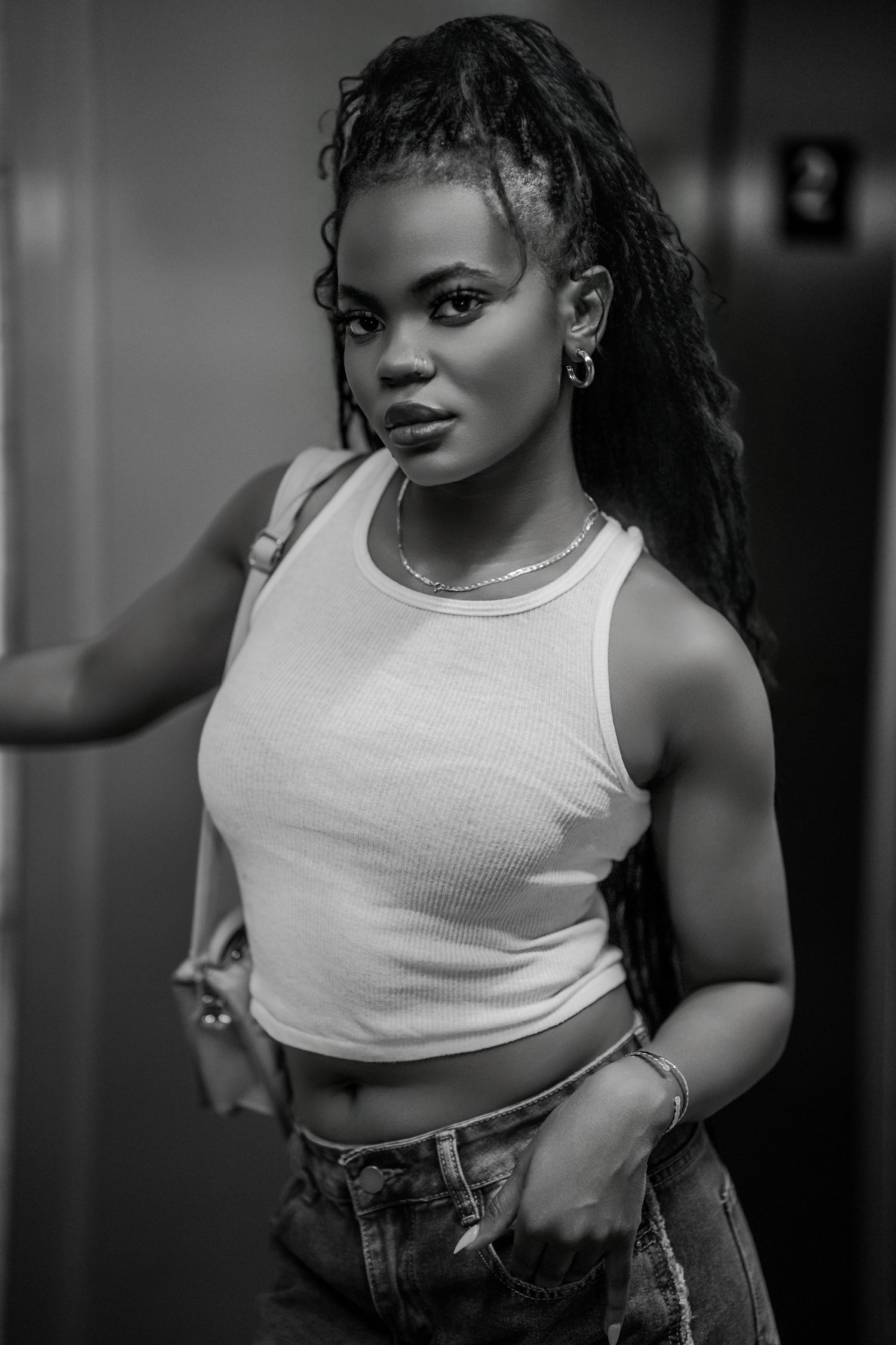 Black and white portrait of a young woman with braids wearing a crop top and jeans, posing confidently indoors.