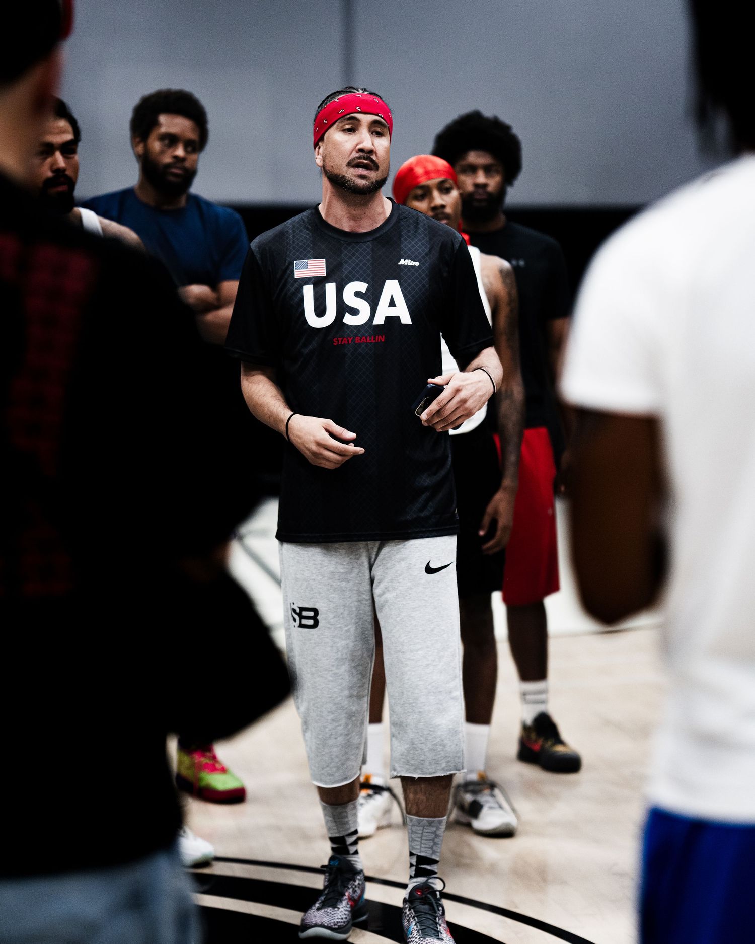 Coach in USA black shirt and red headband addresses a group of basketball players during an indoor training session.