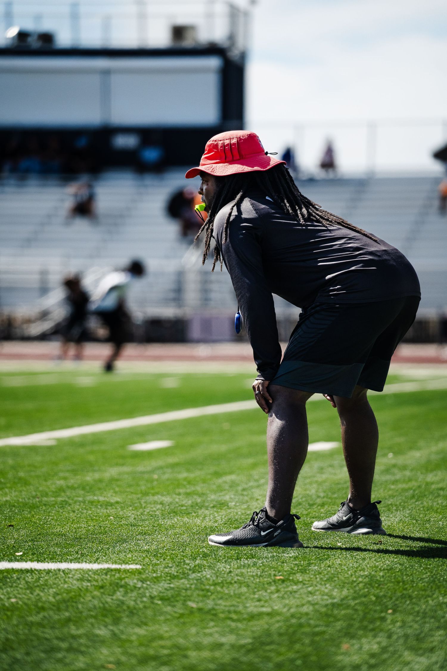 Coach in red bucket hat observes athletes during a football training camp on a green turf field.