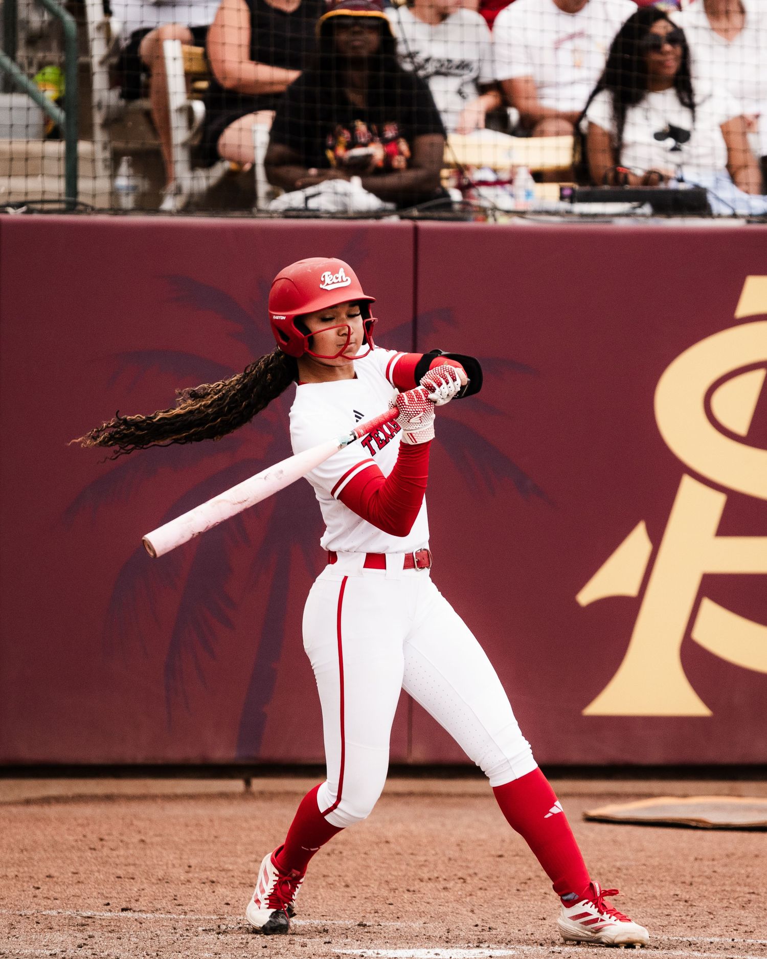 Softball player in red and white uniform swings bat during college game with fans in background.