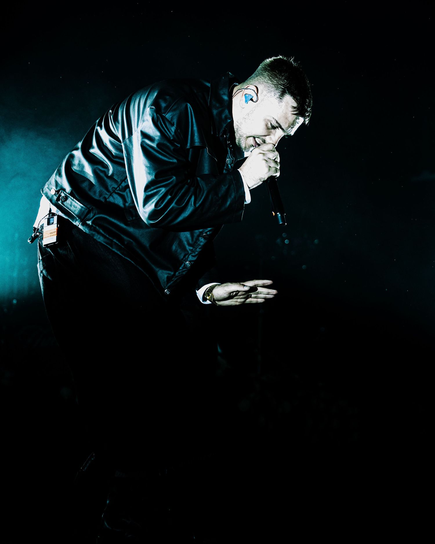 Performer in black jacket bowing on dark stage with dramatic blue lighting during live concert.