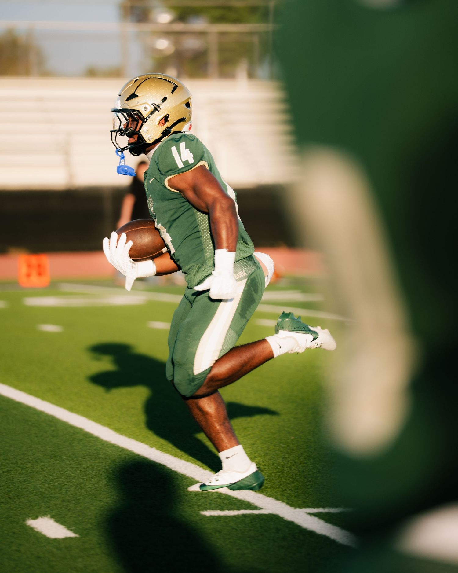 Football player #14 in green and gold uniform runs with the ball on a turf field during a game.