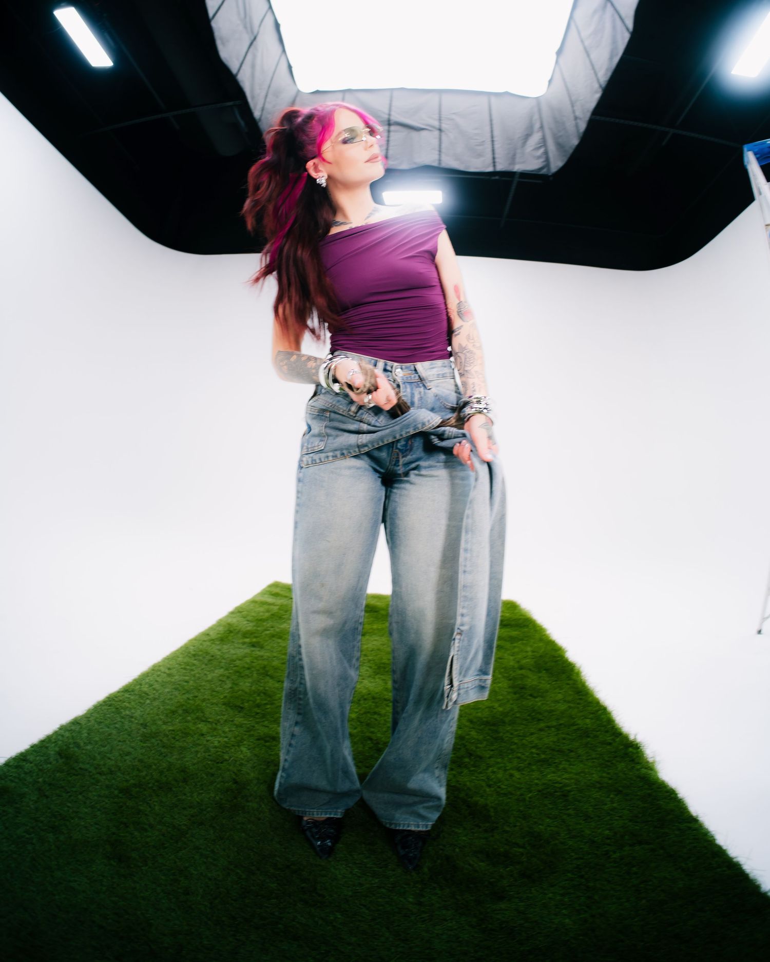 Woman with pink hair in purple top and wide-leg jeans poses in white studio with artificial grass floor.