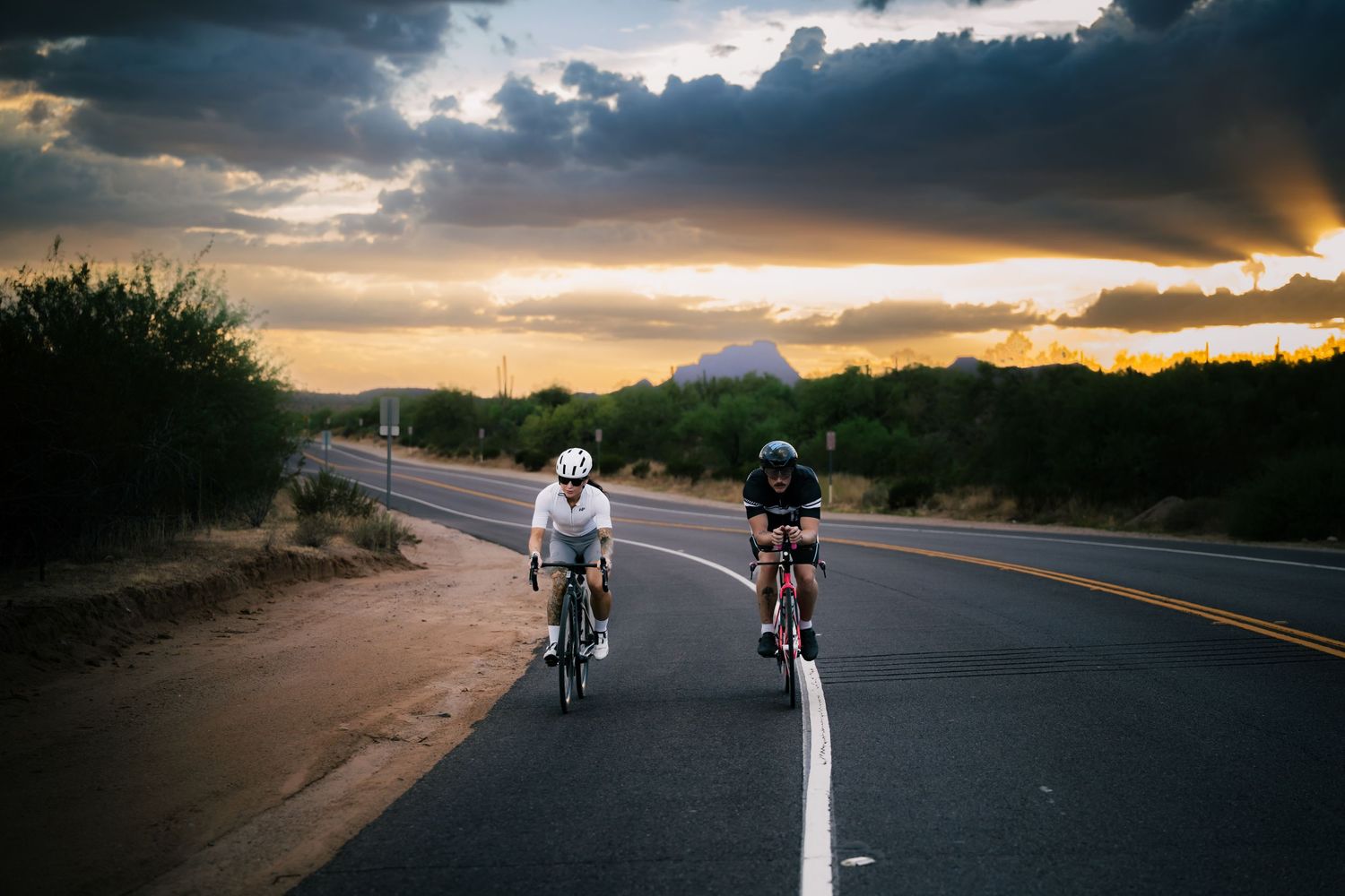 Two cyclists ride road bikes at sunset on a desert highway under dramatic cloudy skies.