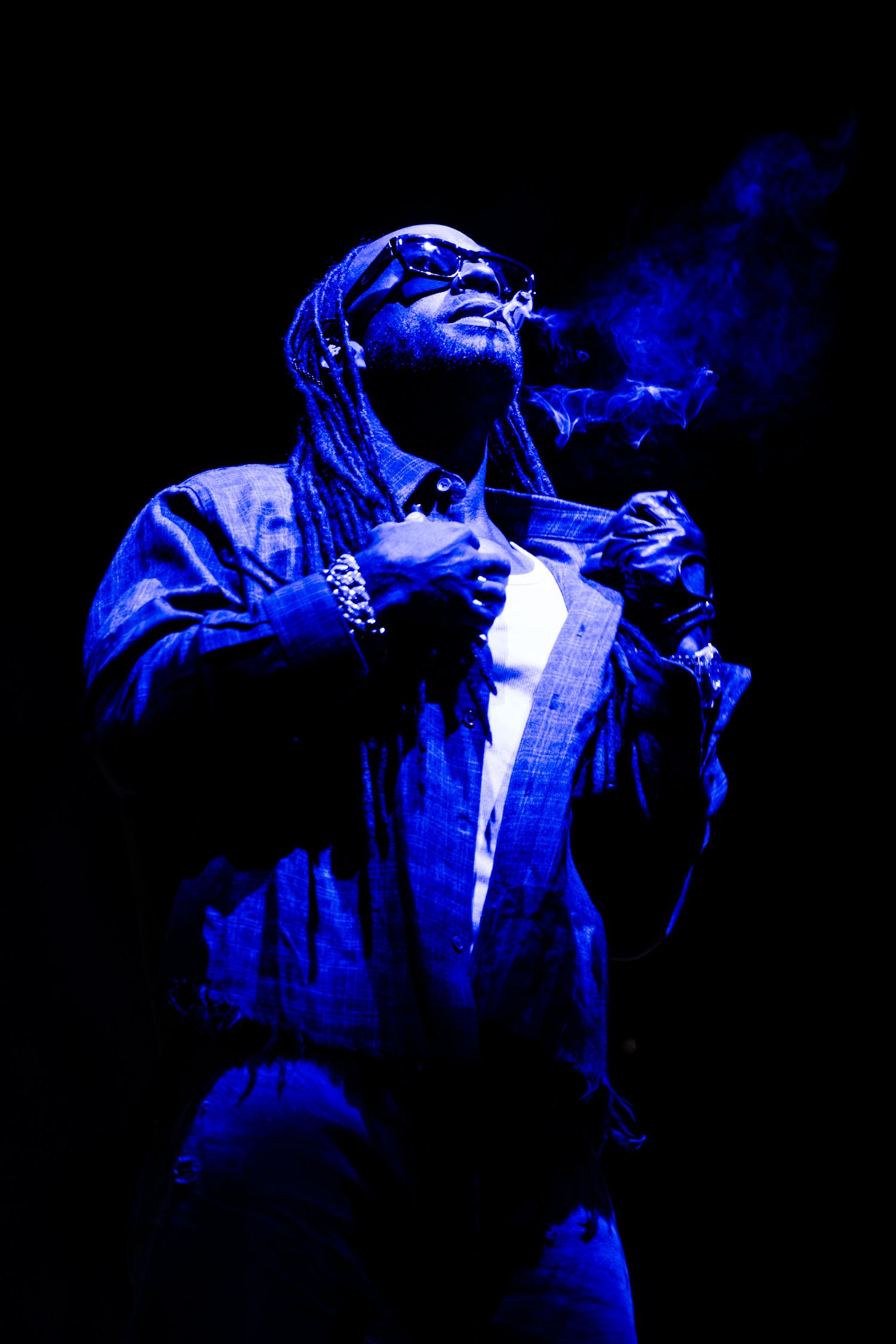 Man with long braids and sunglasses exhales smoke while adjusting his jacket collar, lit in dramatic blue light against a dark background.