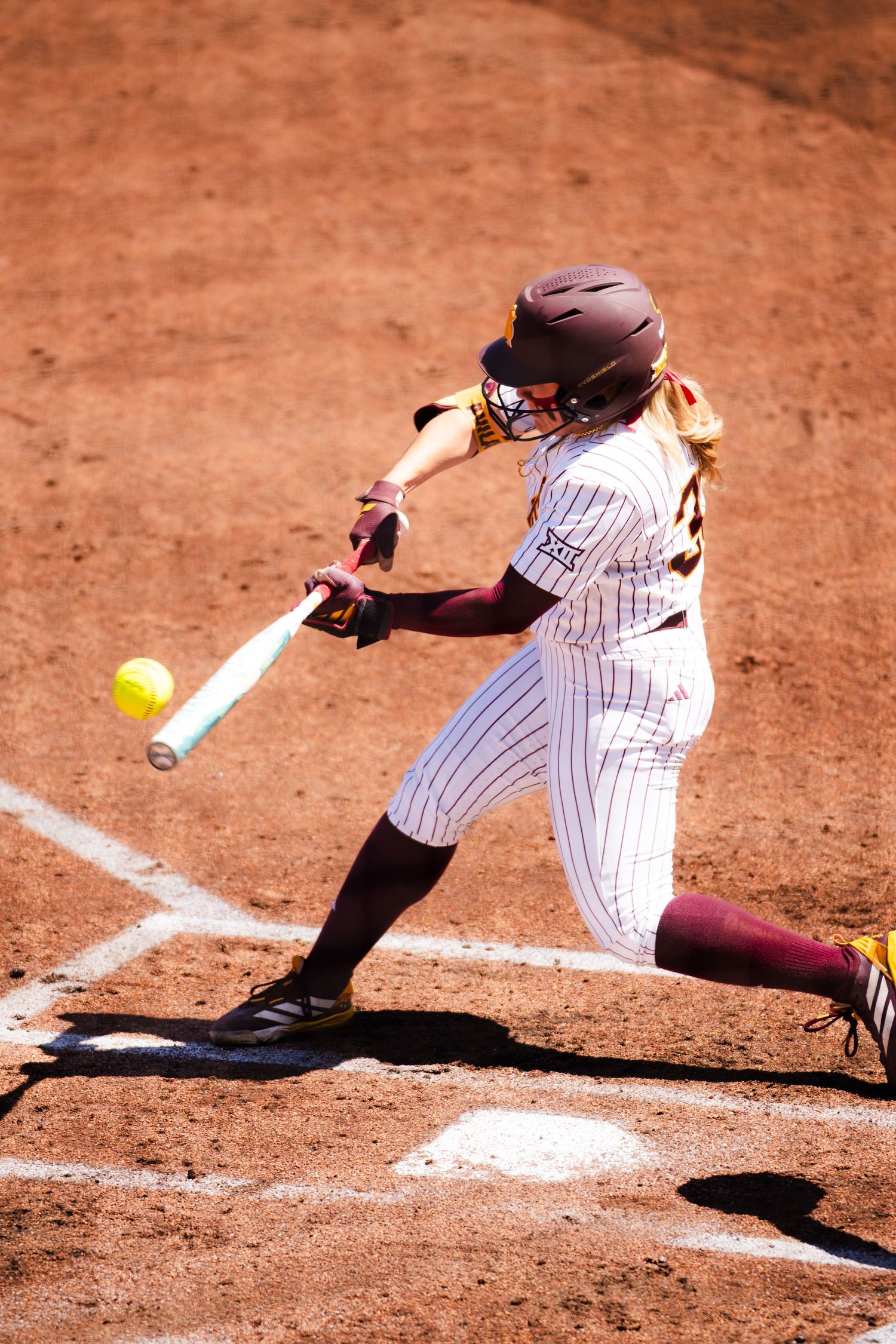 Female softball player in maroon uniform swinging bat at yellow ball during a sunny game on a dirt field.
