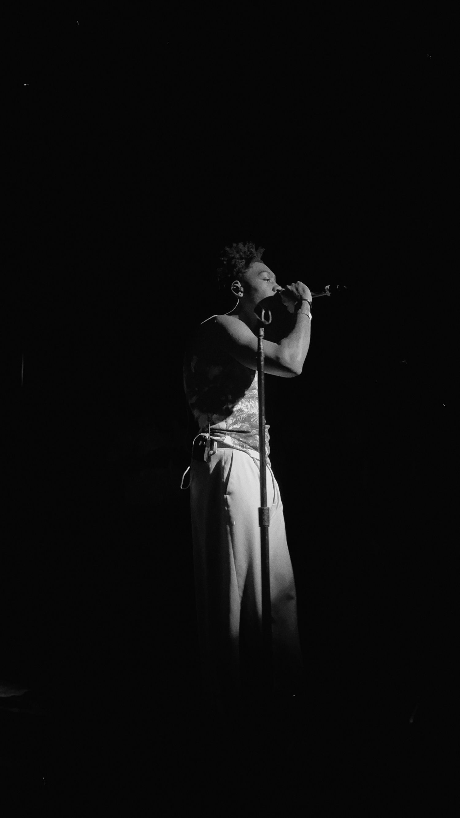 Black and white photo of a singer performing on stage with a microphone stand in dramatic dark lighting.
