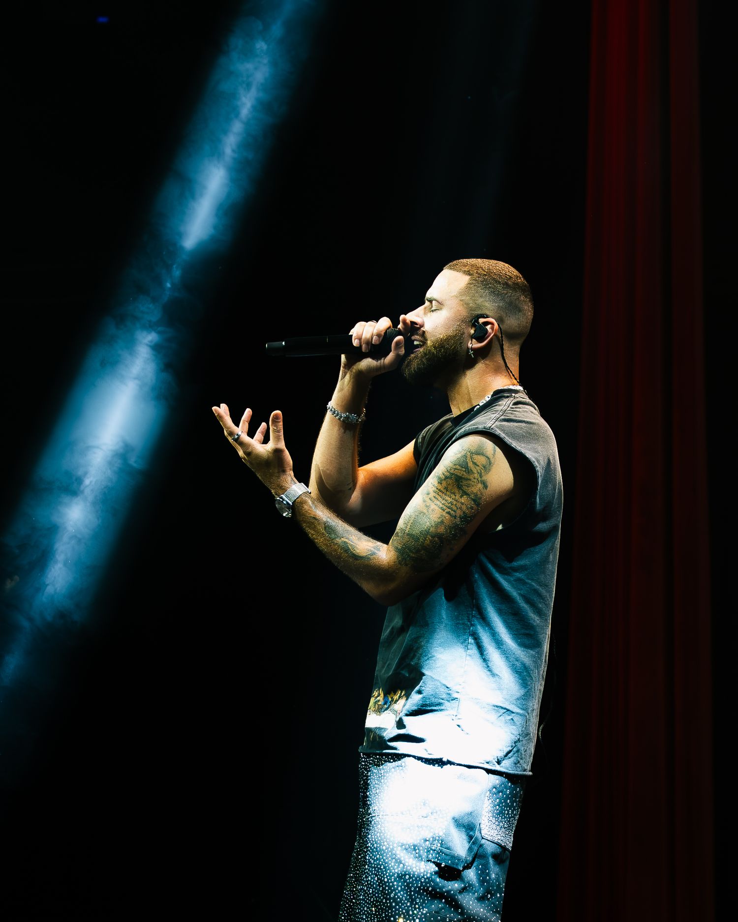 Tattooed male singer performing on stage with microphone under dramatic blue spotlight in dark concert venue.
