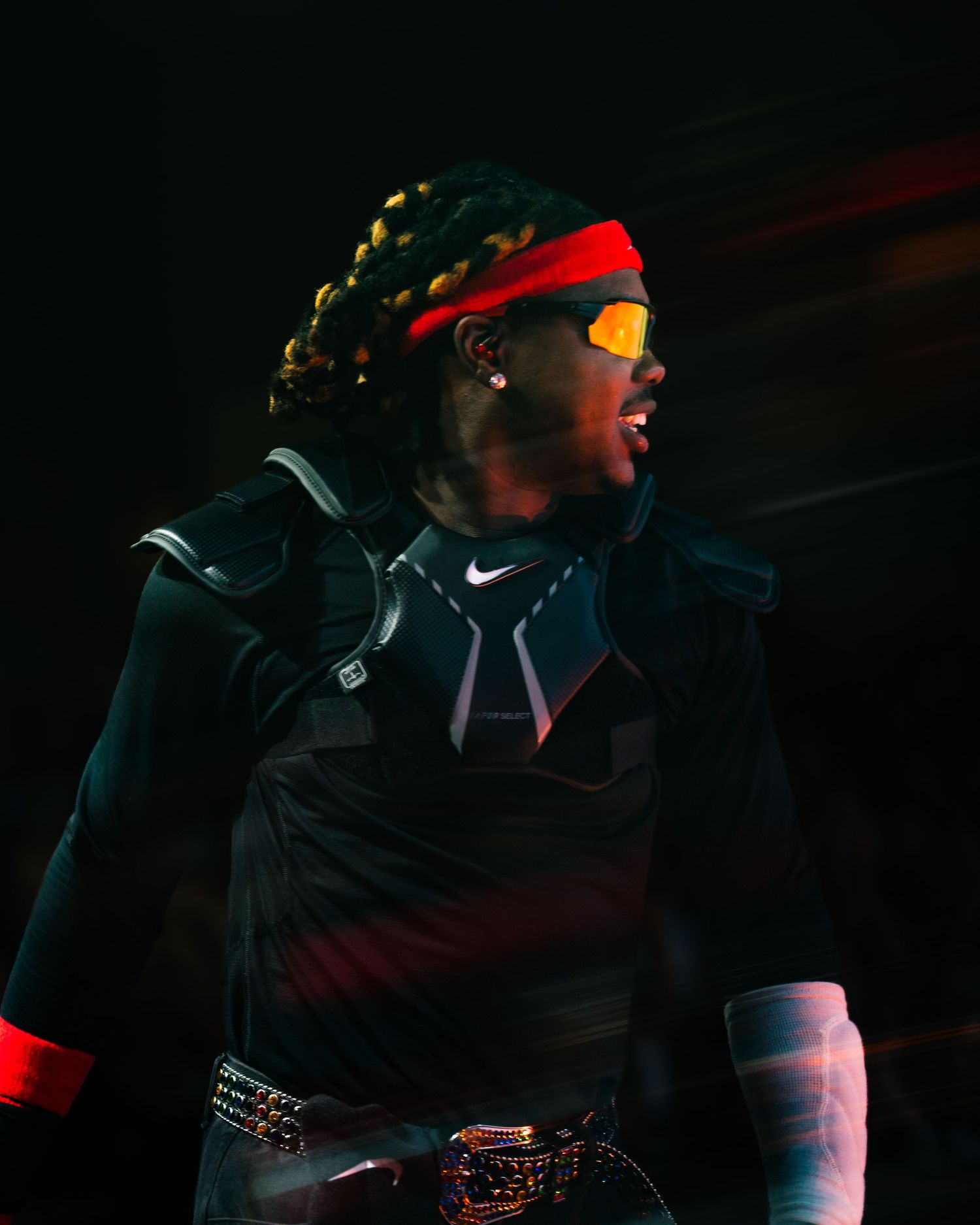 Athlete in Nike gear with braids, red headband, and orange sunglasses performing under dramatic stage lighting.