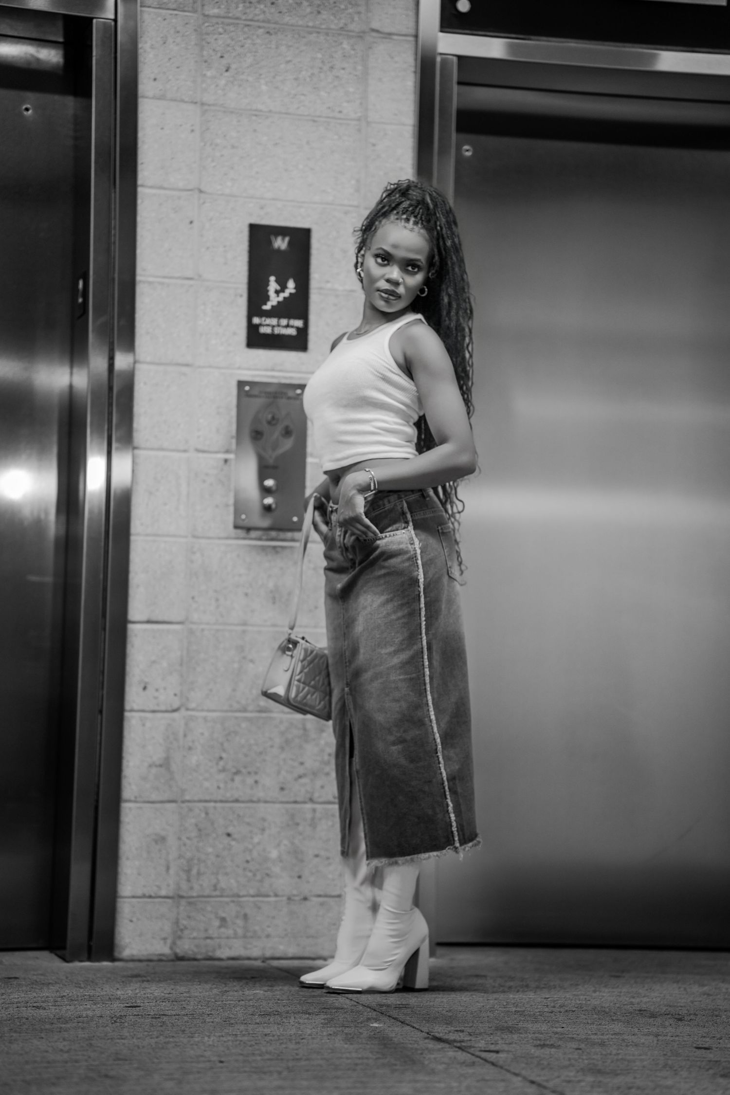 Black and white photo of a stylish young woman in white crop top, denim midi skirt, and white boots near elevators.