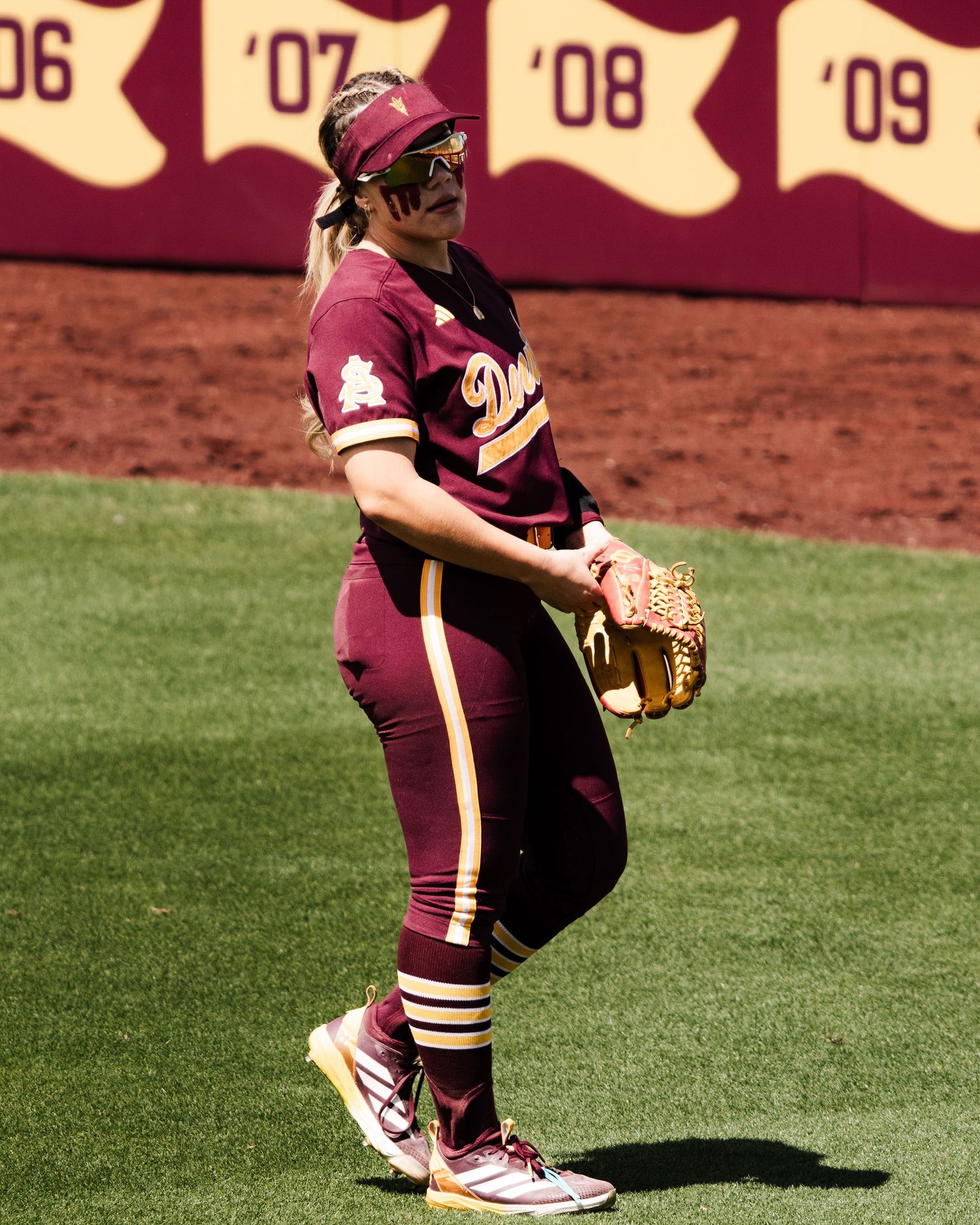 Female softball player in maroon uniform with glove, standing on field with numbered banners in background.