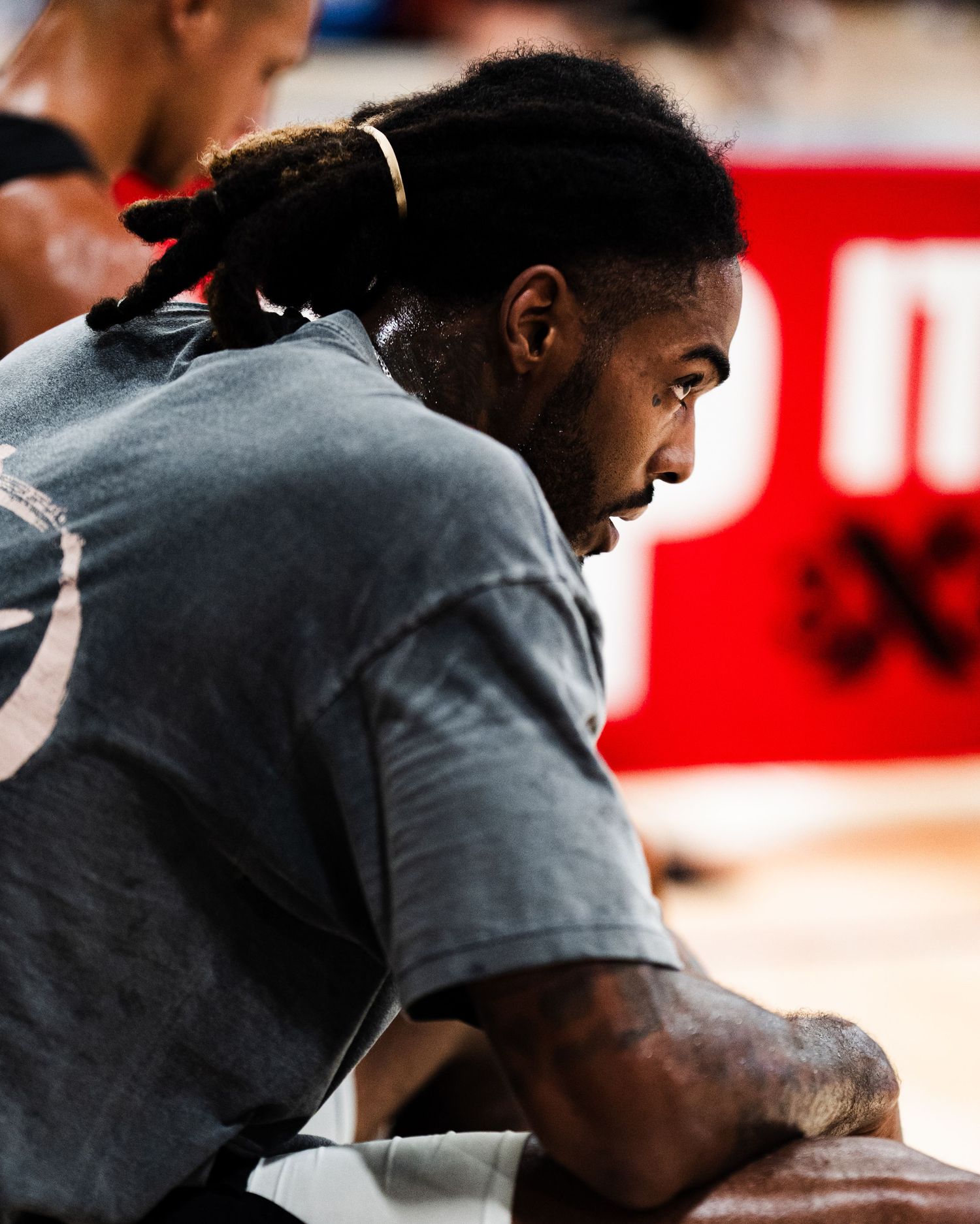 A man with braided hair in a gray t-shirt sits courtside at a basketball event, viewed from behind.