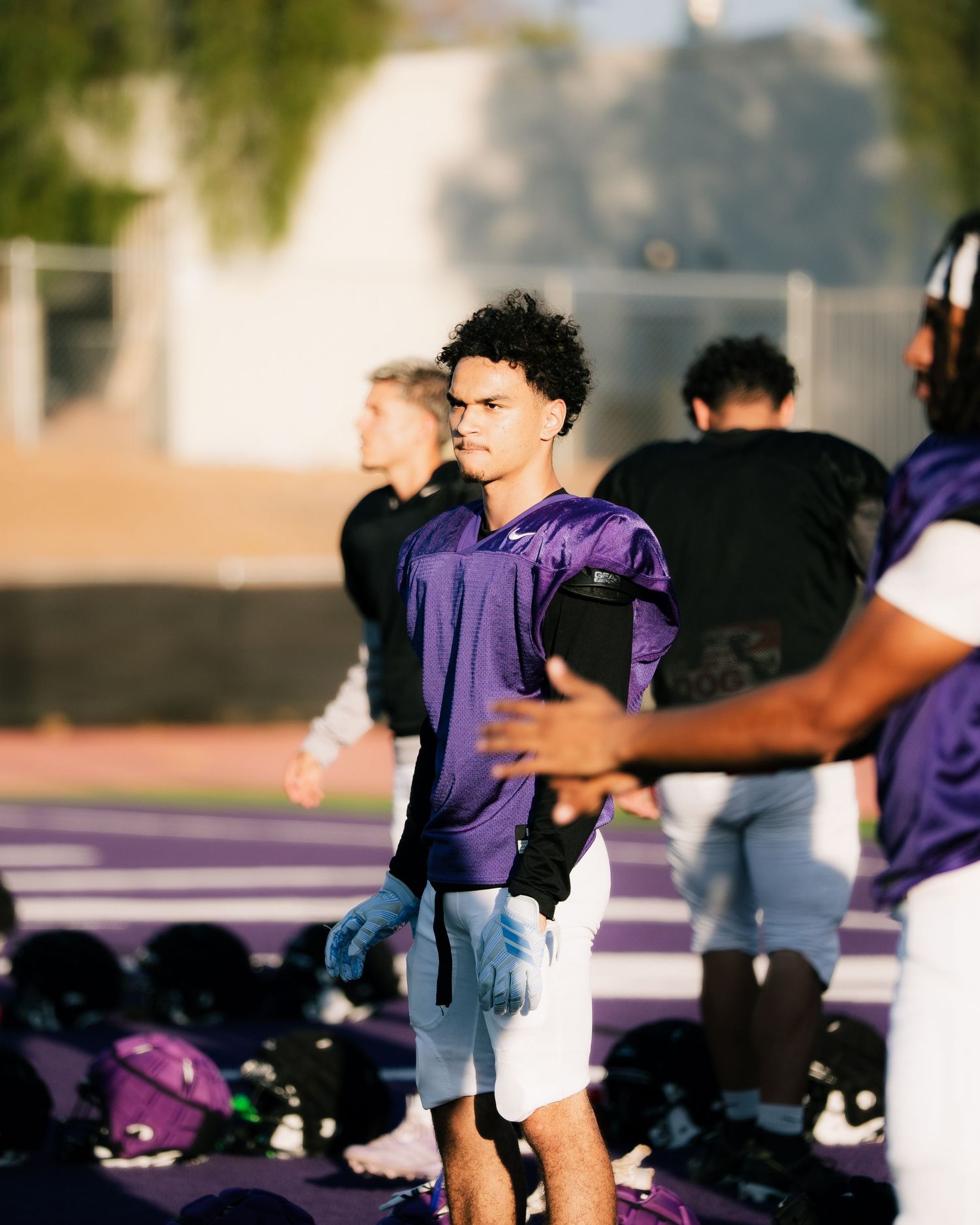 Football players in purple jerseys practice drills on a purple turf field during an outdoor team training session.