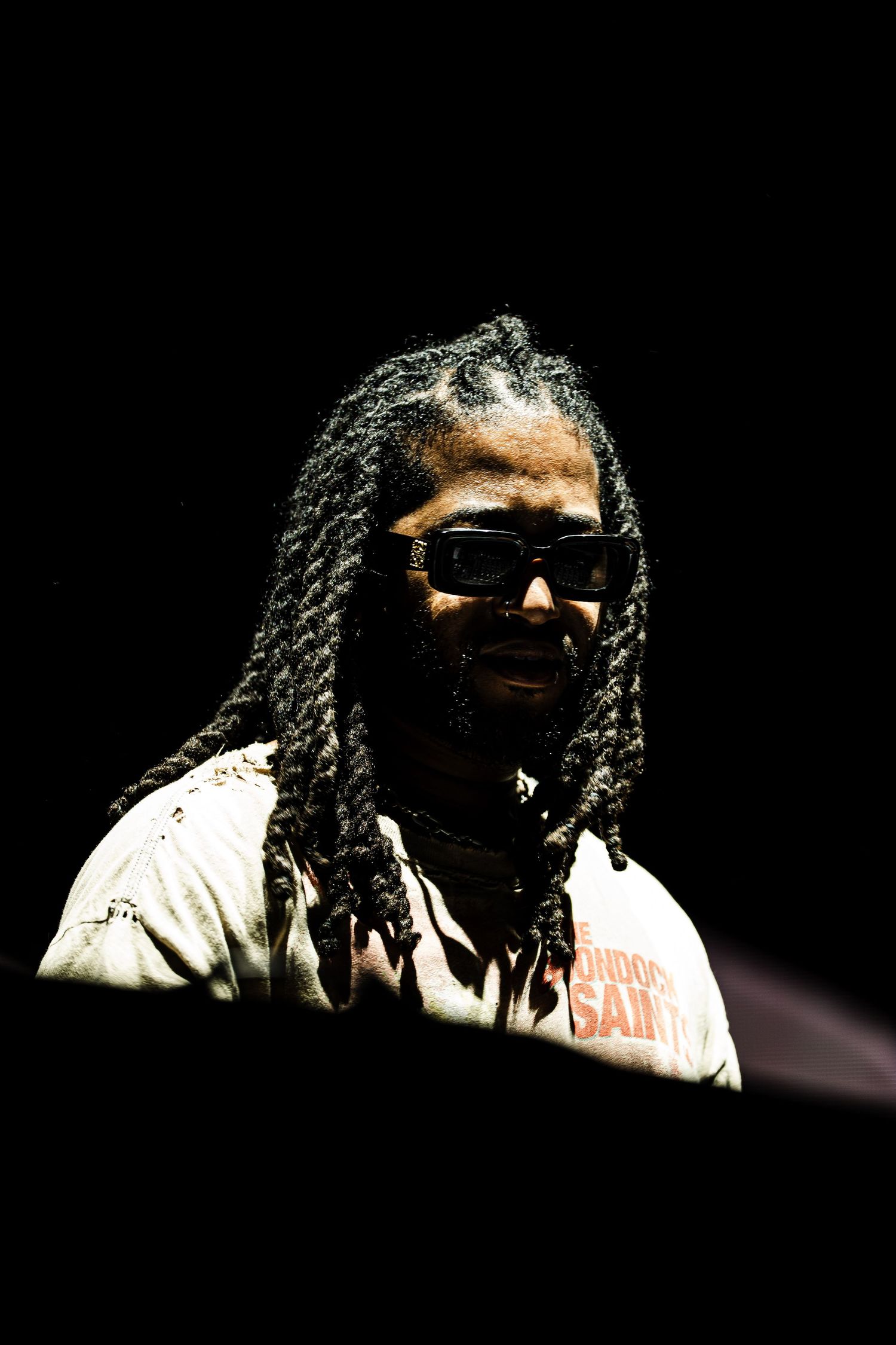 Man with long twisted locs and dark sunglasses wearing a distressed light graphic t-shirt, lit dramatically against a black background.