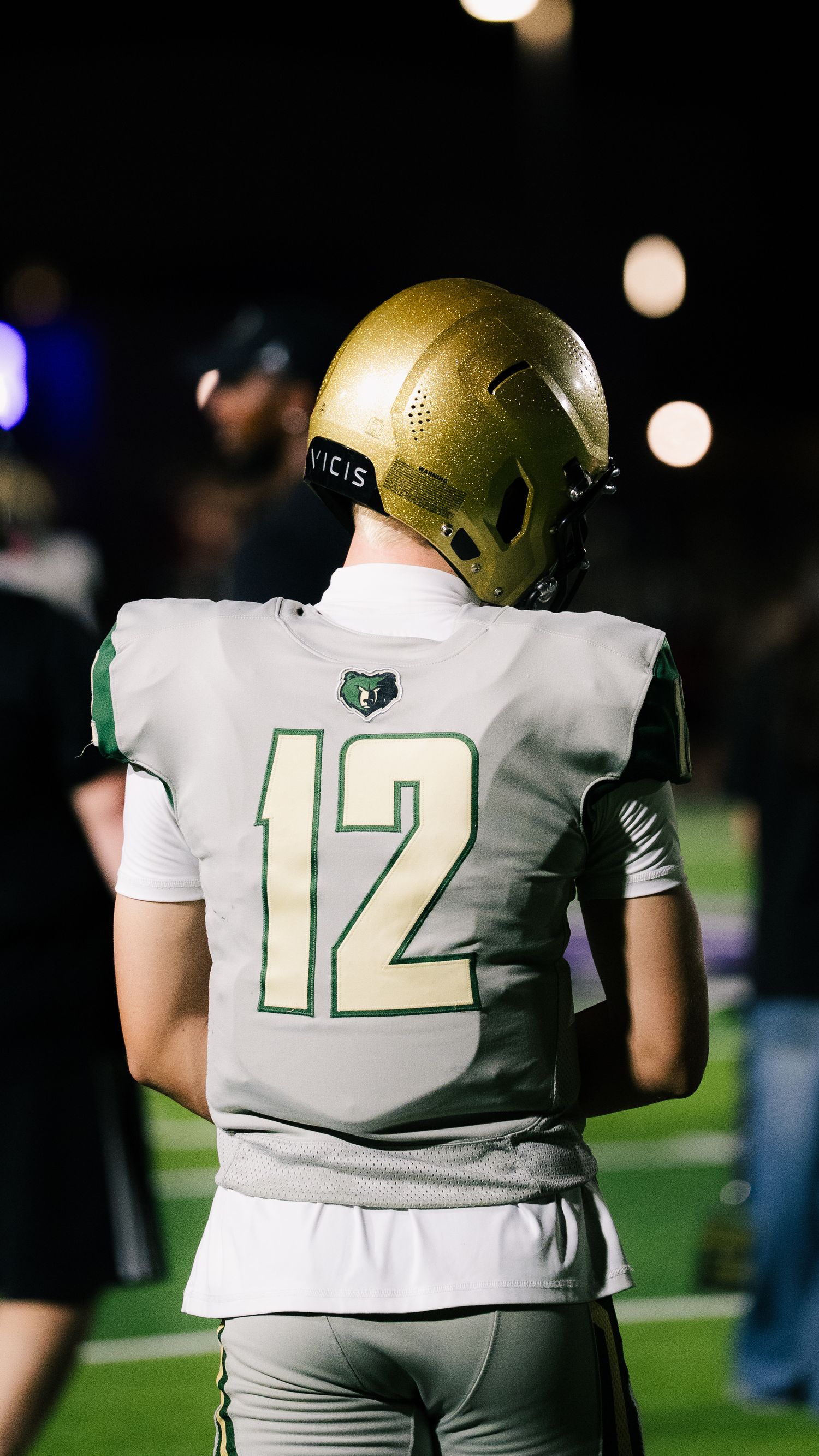 Football player #12 in white and green jersey with gold helmet stands on sideline during night game.