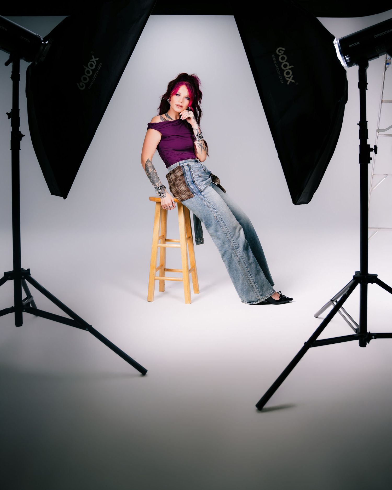 Woman with pink hair poses on stool in purple top and wide-leg jeans, surrounded by Godox studio lights.