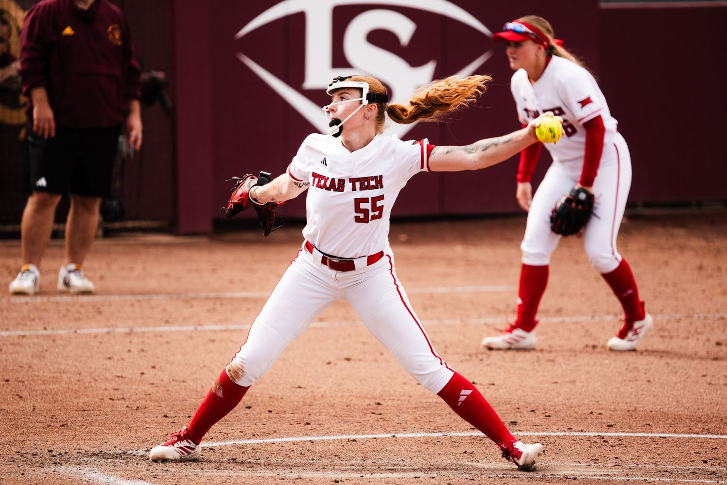 Texas Tech softball pitcher #55 winds up for a pitch during a college softball game on a red dirt field.