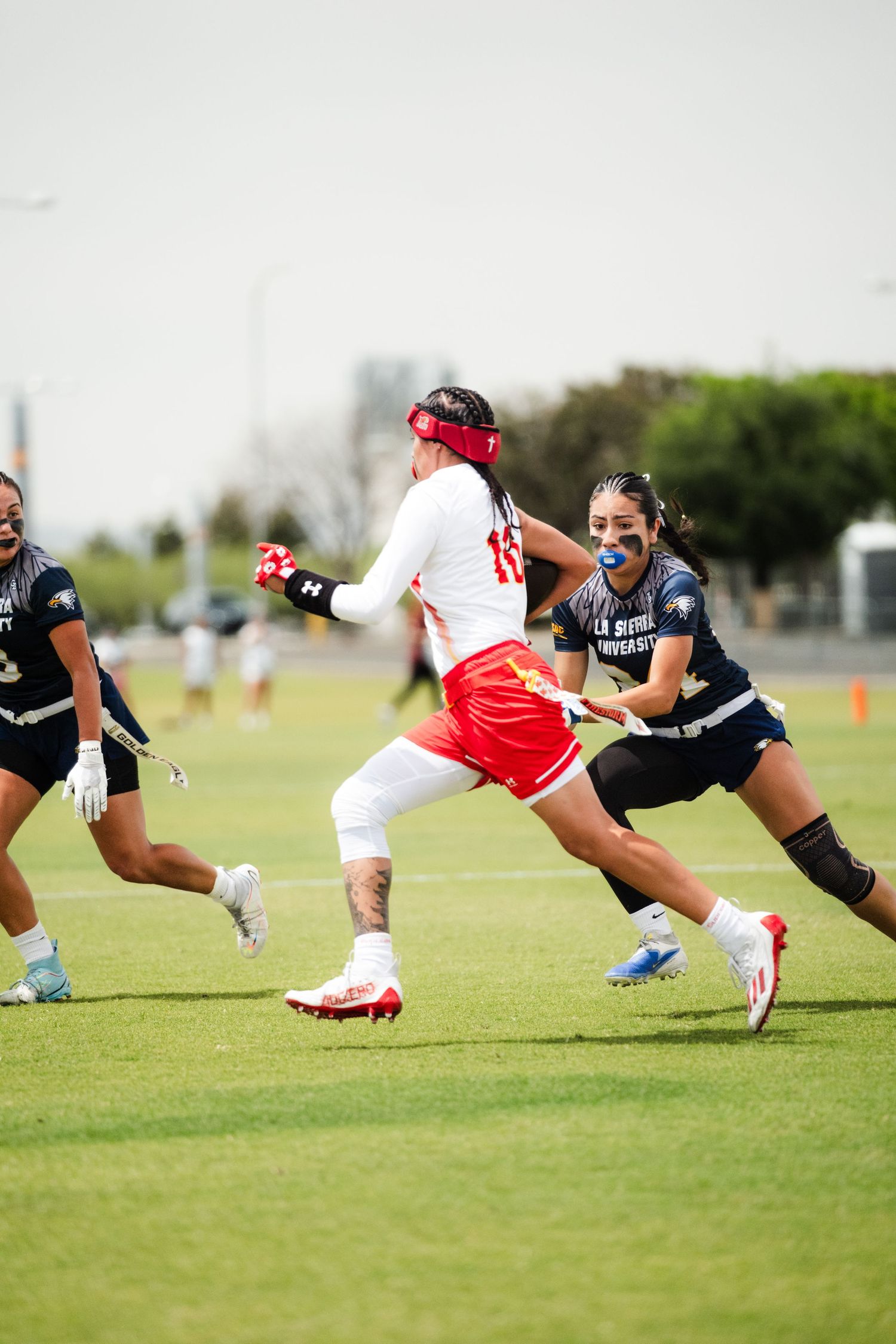 Women's flag football players compete on grass field, defender reaching for flag on red-uniformed ball carrier.