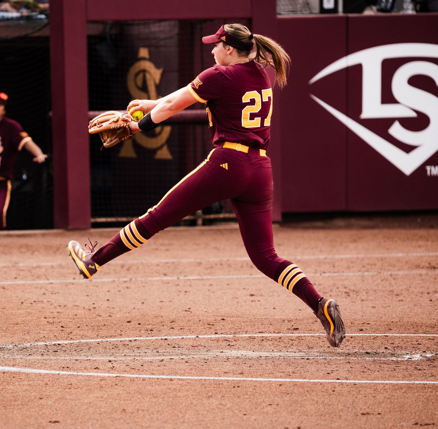 College softball pitcher #27 in maroon uniform mid-windup delivery on dirt mound during game.