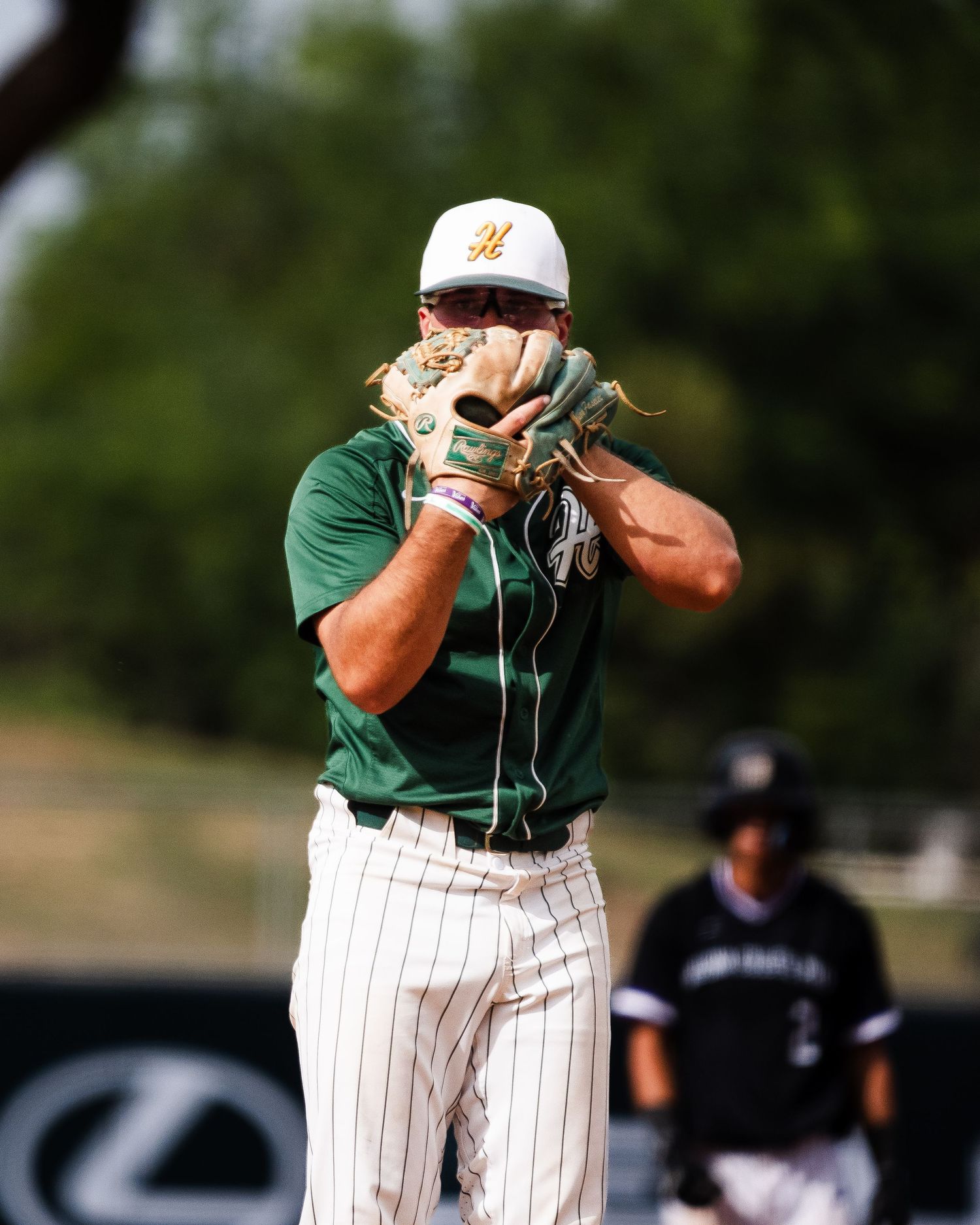 Baseball pitcher in green uniform and white cap winds up to throw during a sunny outdoor game.