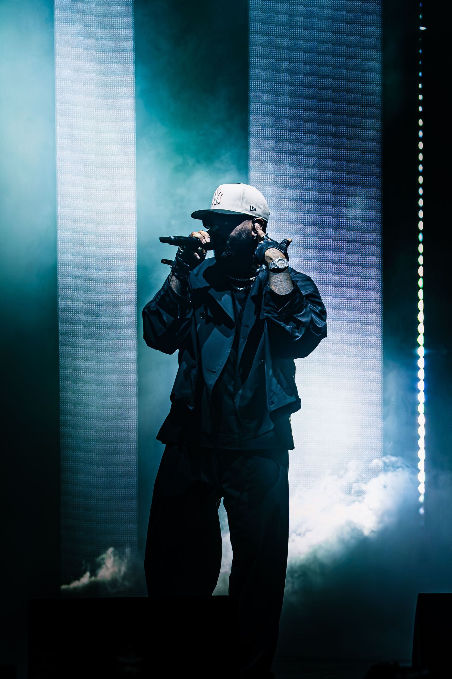 Performer in a dark jacket, white cap and face covering raps into a microphone on a smoky stage with glowing teal lights.