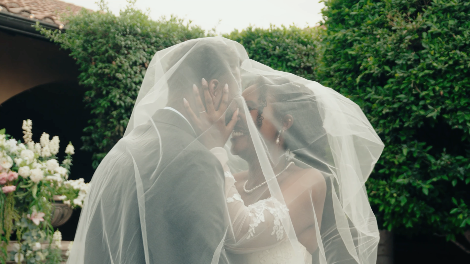 Bride and groom sharing a romantic kiss under a flowing white wedding veil outdoors surrounded by lush greenery.