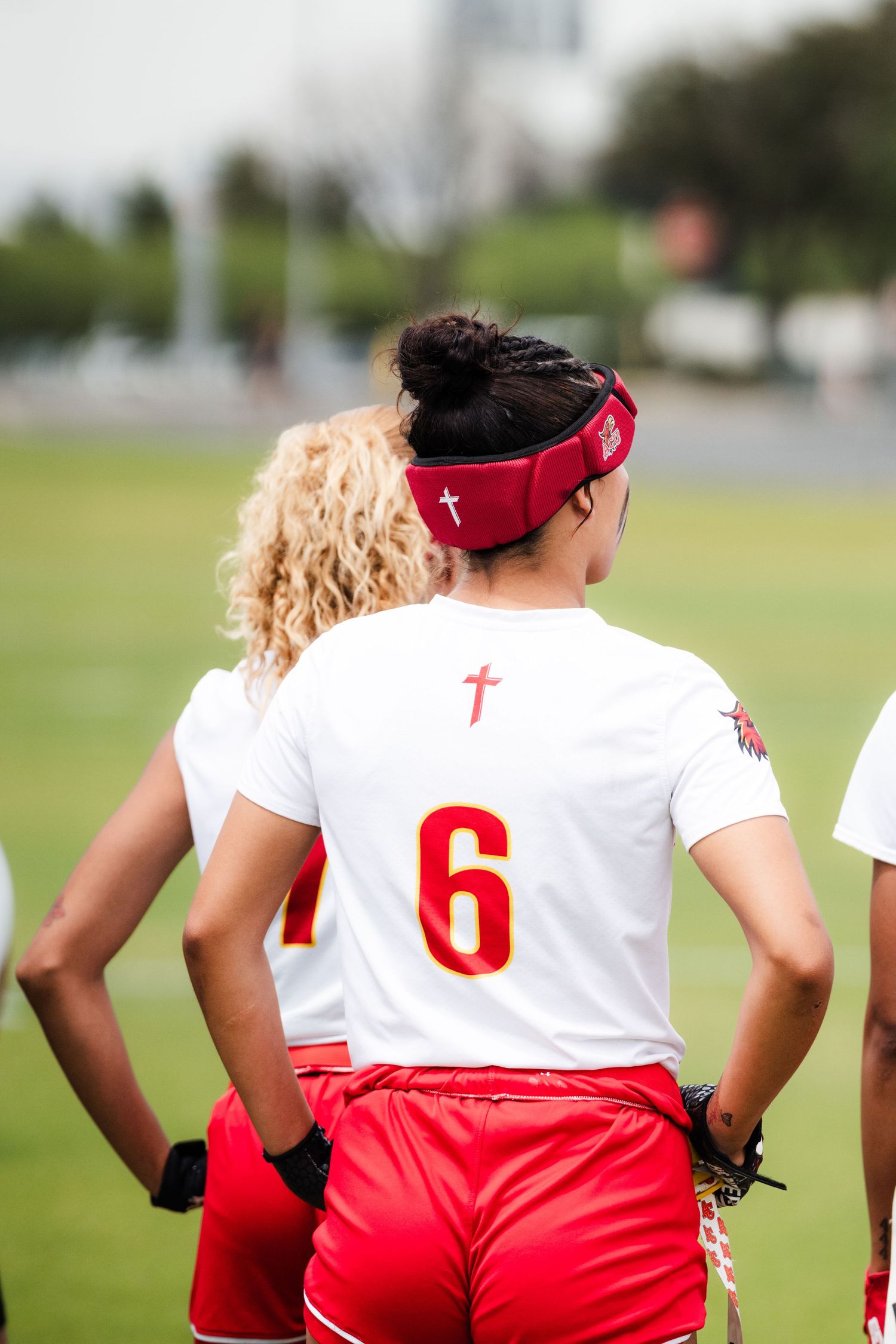 Female flag football player wearing number 6 red and white uniform with cross logo stands on green field.