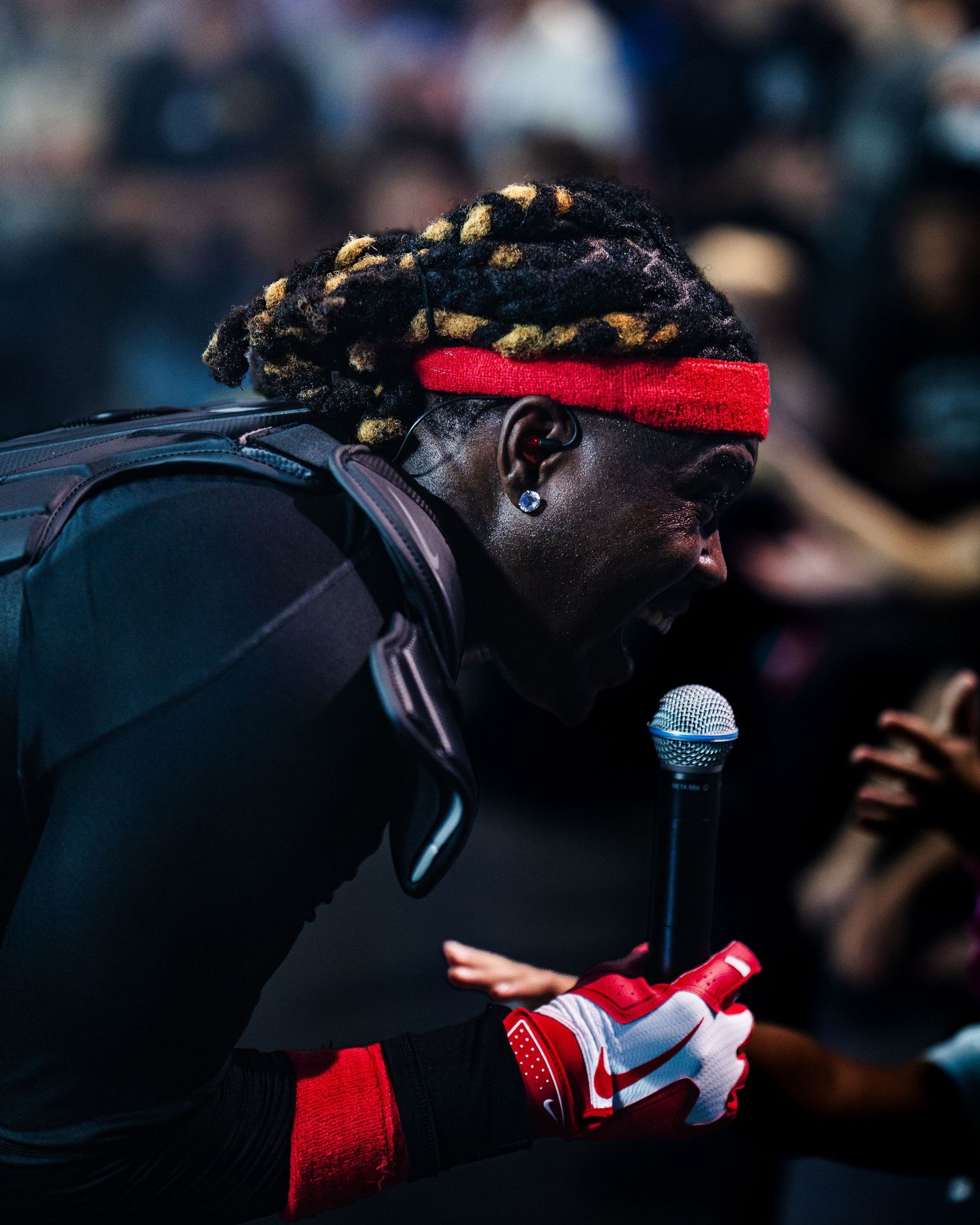 Performer with gold-tipped dreadlocks, red headband, and red Nike gloves rapping into a microphone at a live concert.