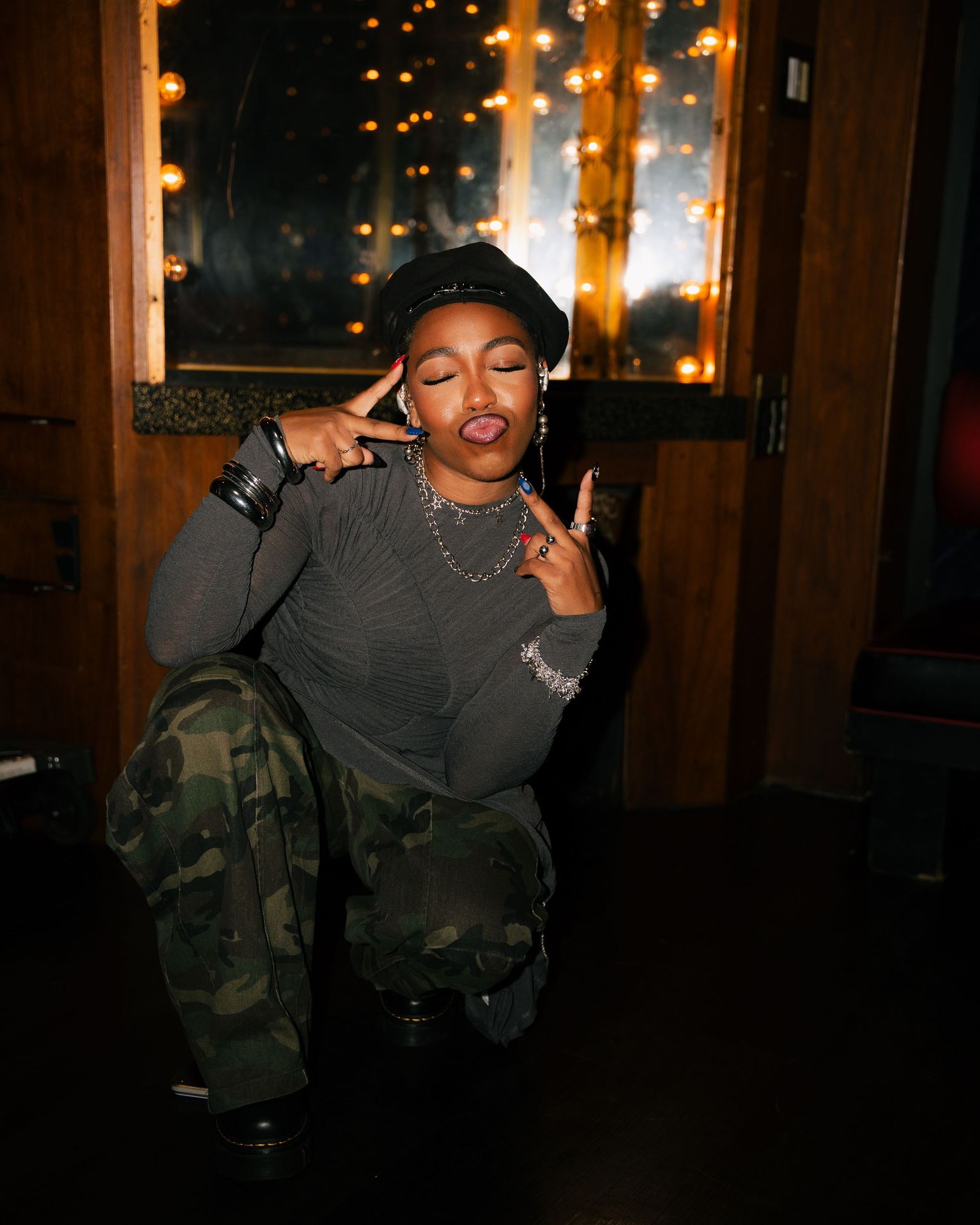 Young woman in camo pants and black cap posing with peace signs indoors.