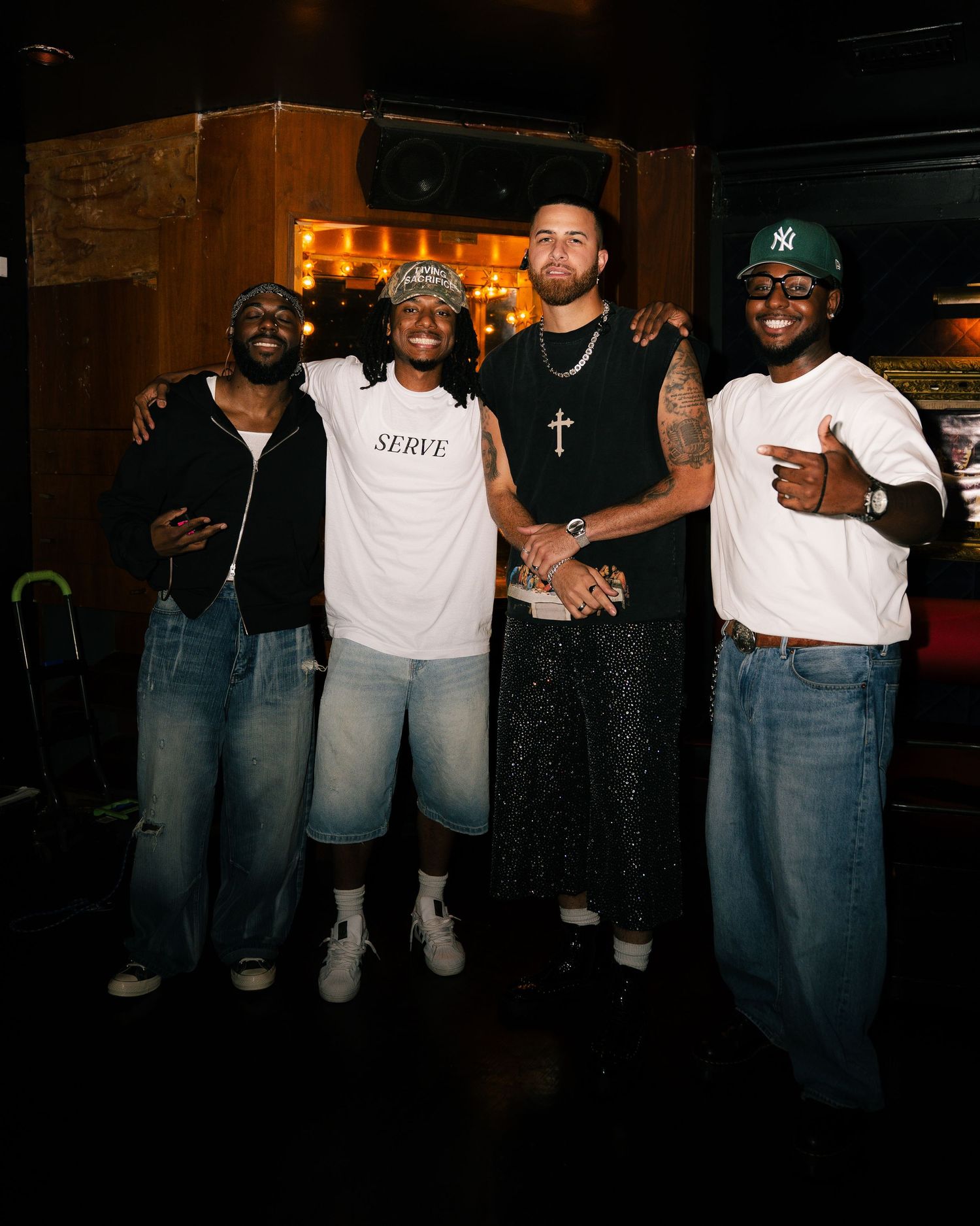 Four men posing together smiling in a dimly lit venue, one wearing a white SERVE shirt and another in a NY cap.