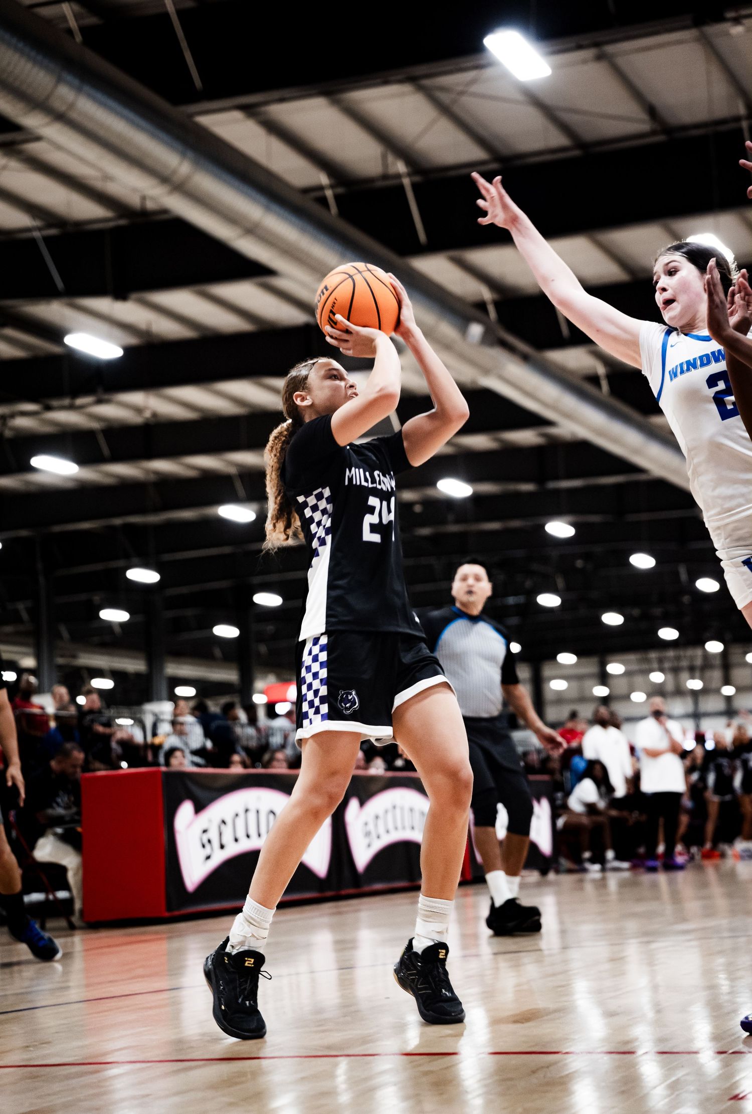 Female basketball player #24 in black Millennium jersey attempts a jump shot during an indoor youth basketball game.