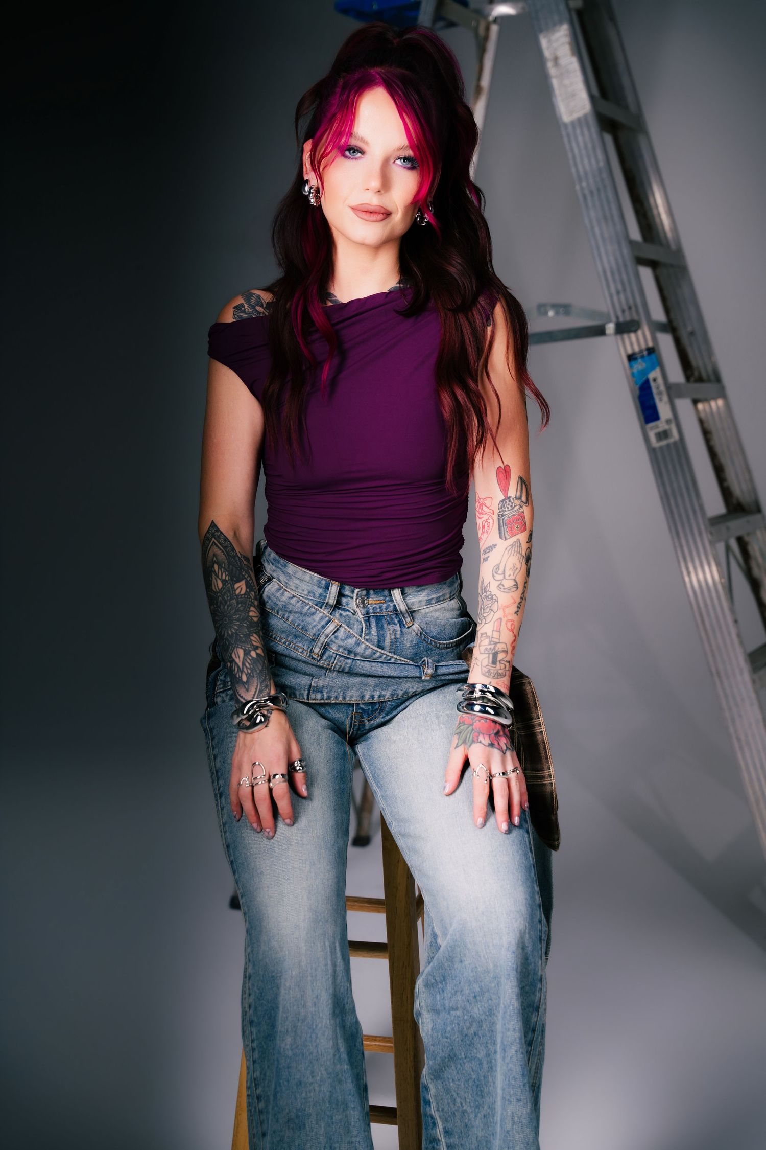 Young woman with pink-tipped hair and tattoos wearing purple top and jeans, posing near a ladder.