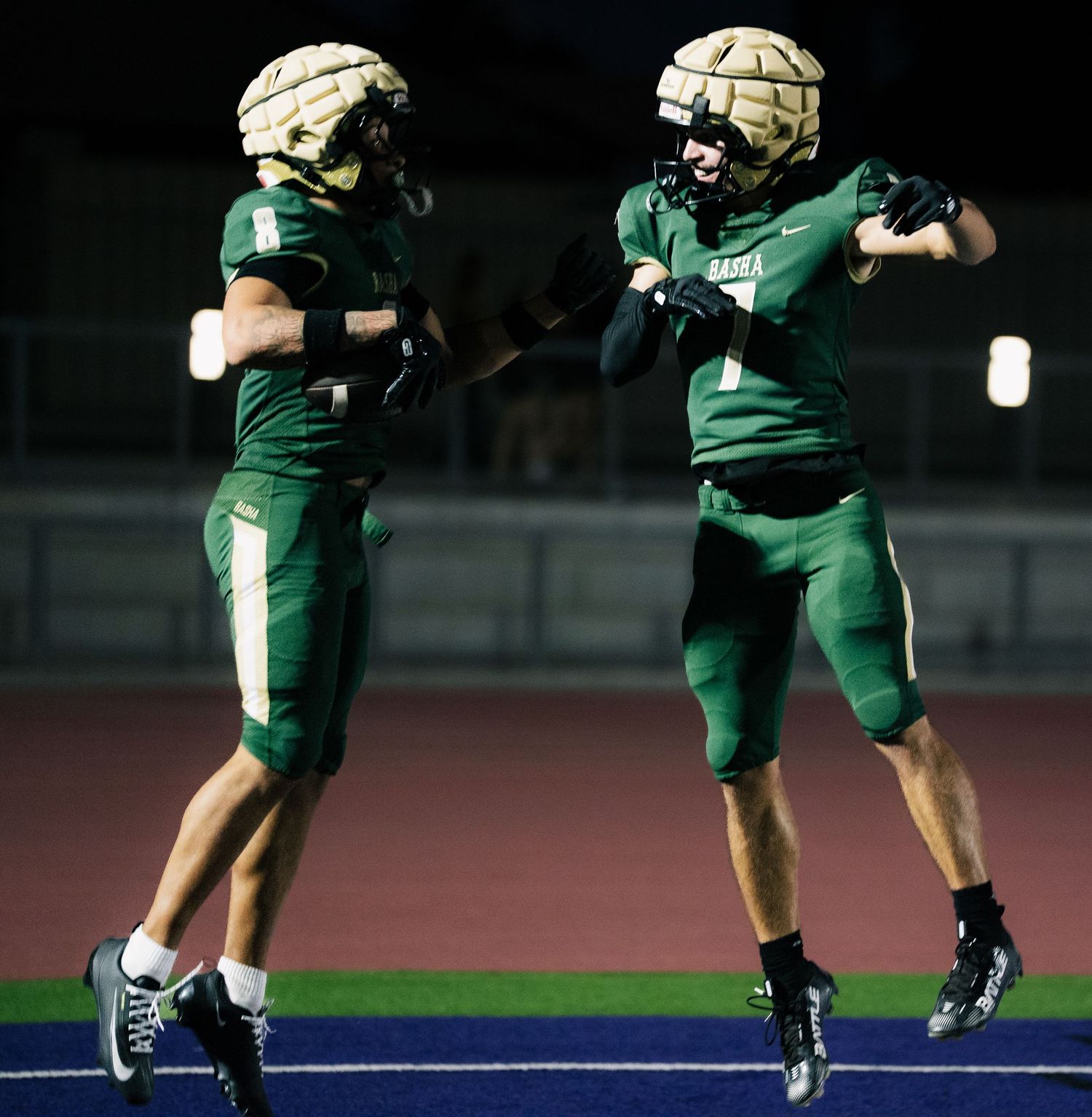 Two football players in green and gold uniforms celebrate on the field during a night game.