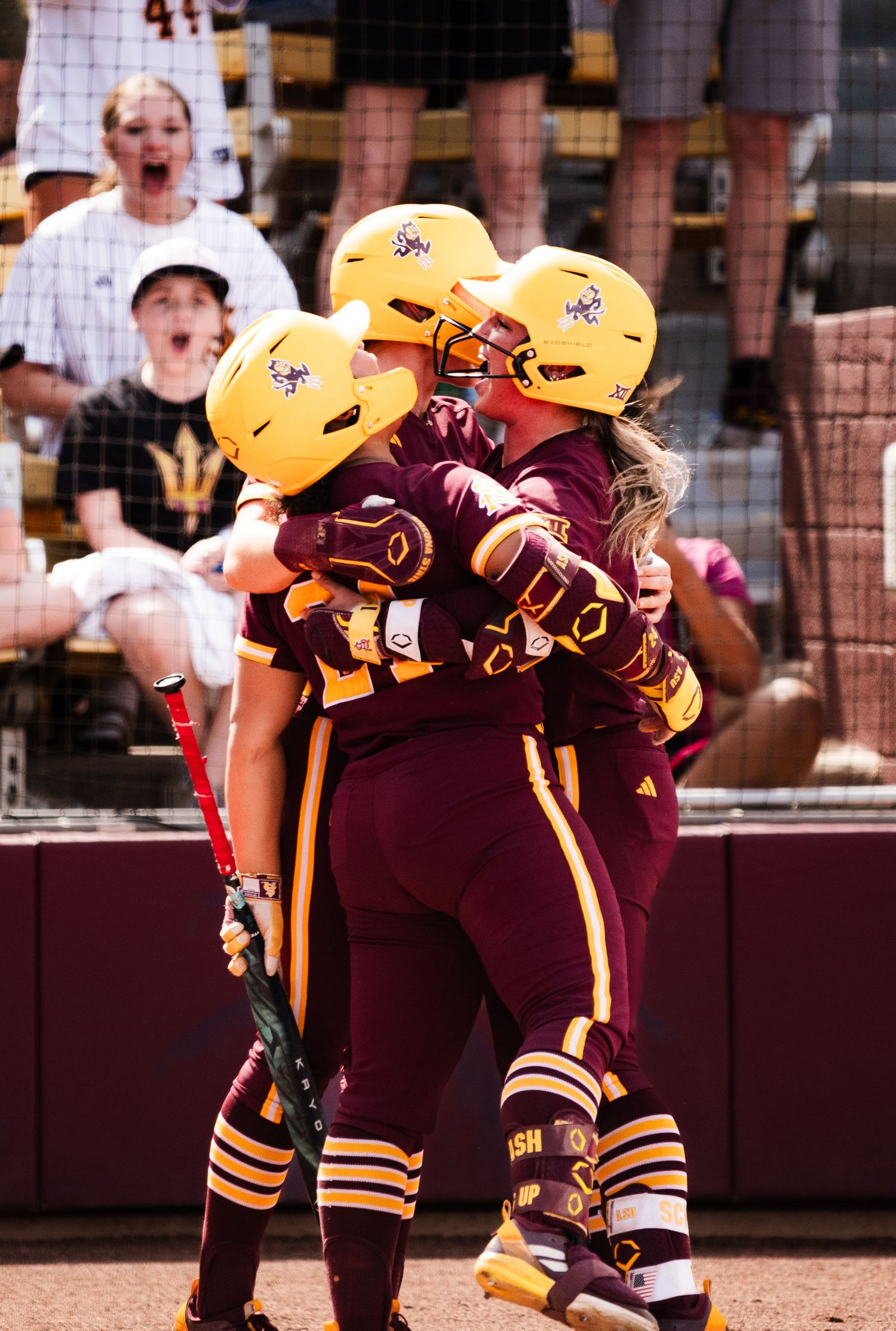 Arizona State Sun Devils softball players celebrate in maroon and gold uniforms and helmets after a big play.