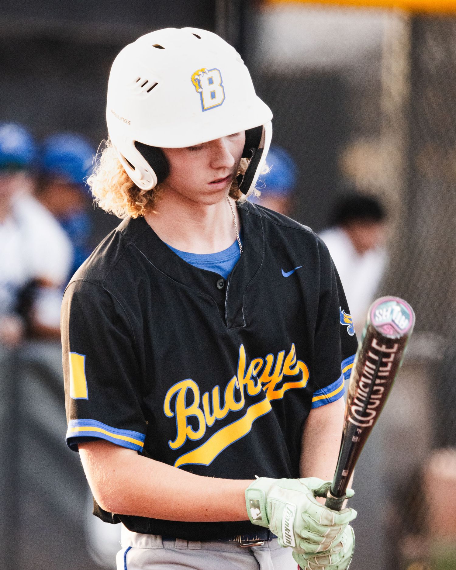 Young baseball player in Buckeyes black jersey and white helmet holds a Louisville Slugger bat during a game.