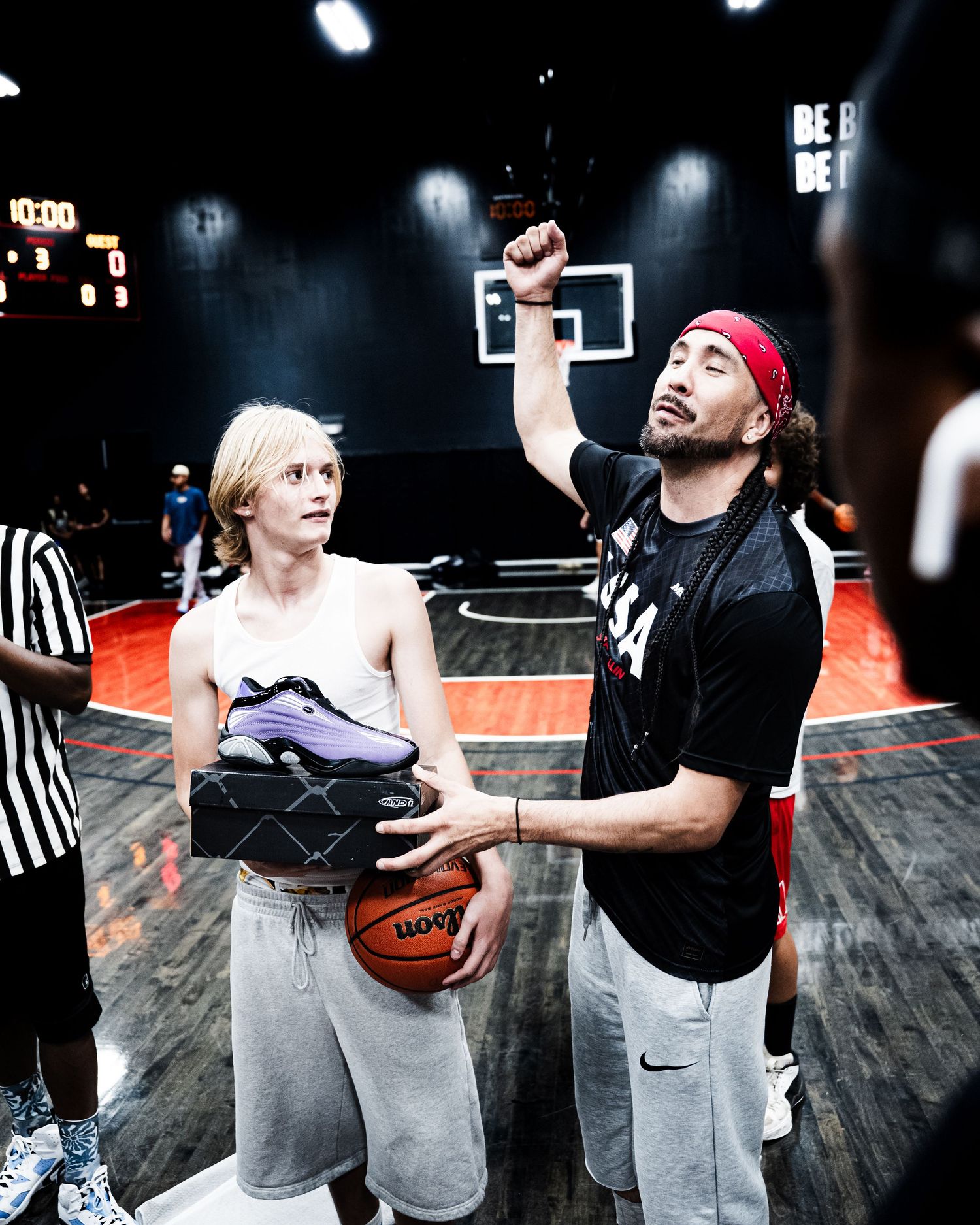 A basketball coach holds up a sneaker while instructing a young player on an indoor court.